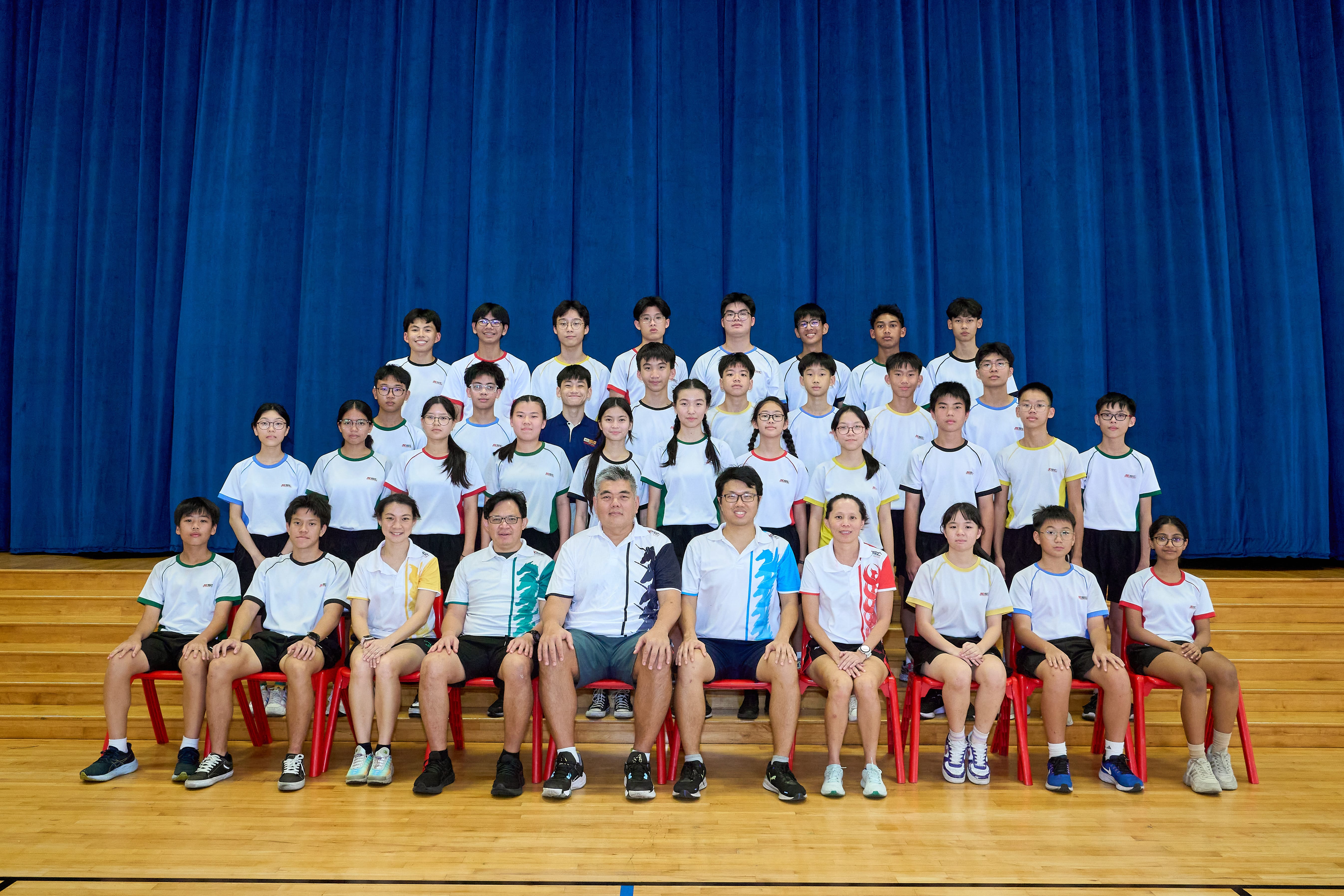Group photo of students and teachers in white uniforms seated and standing before blue curtains on a stage
