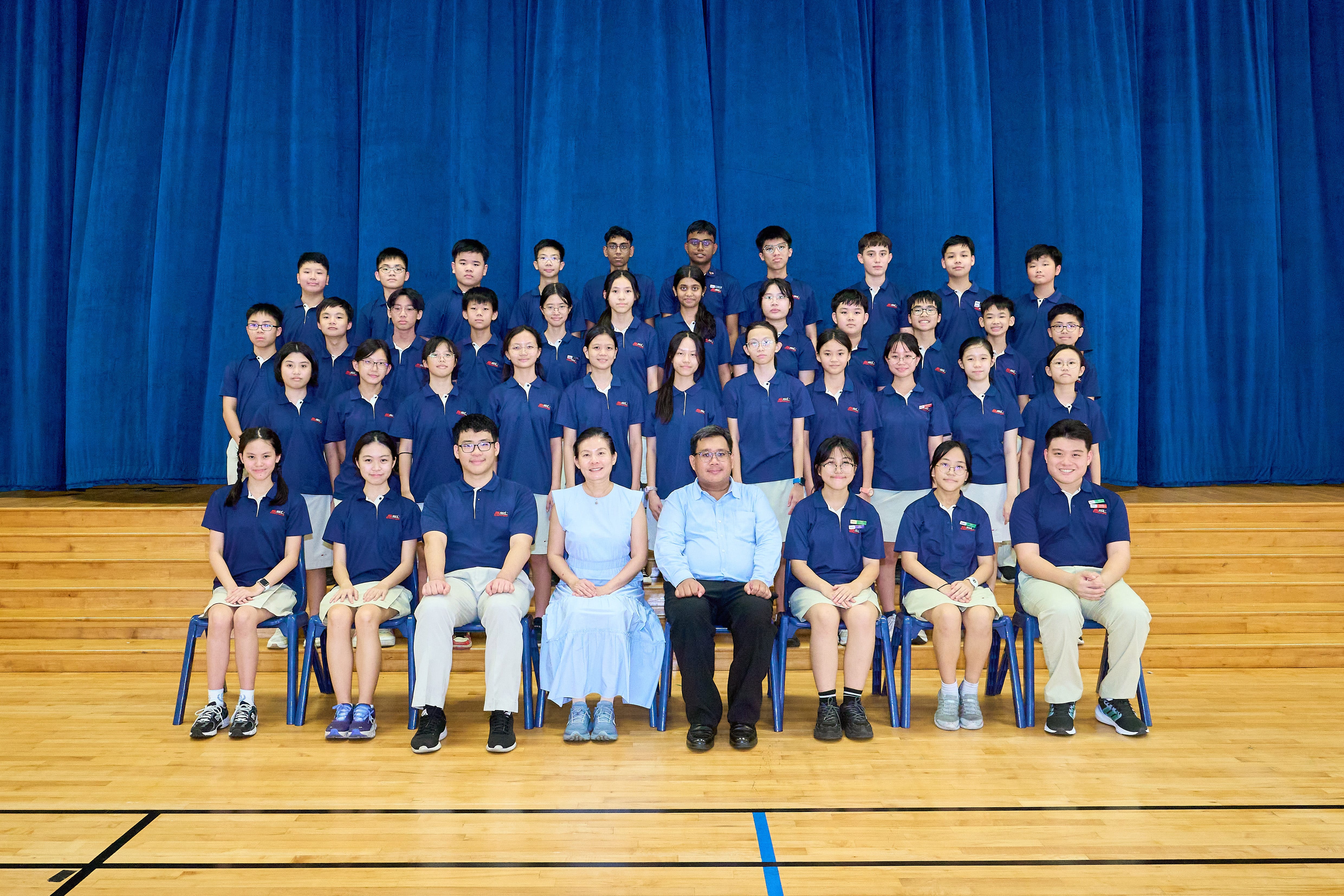 Group of students in navy uniforms posed in rows on a gym stage with blue curtain backdrop.