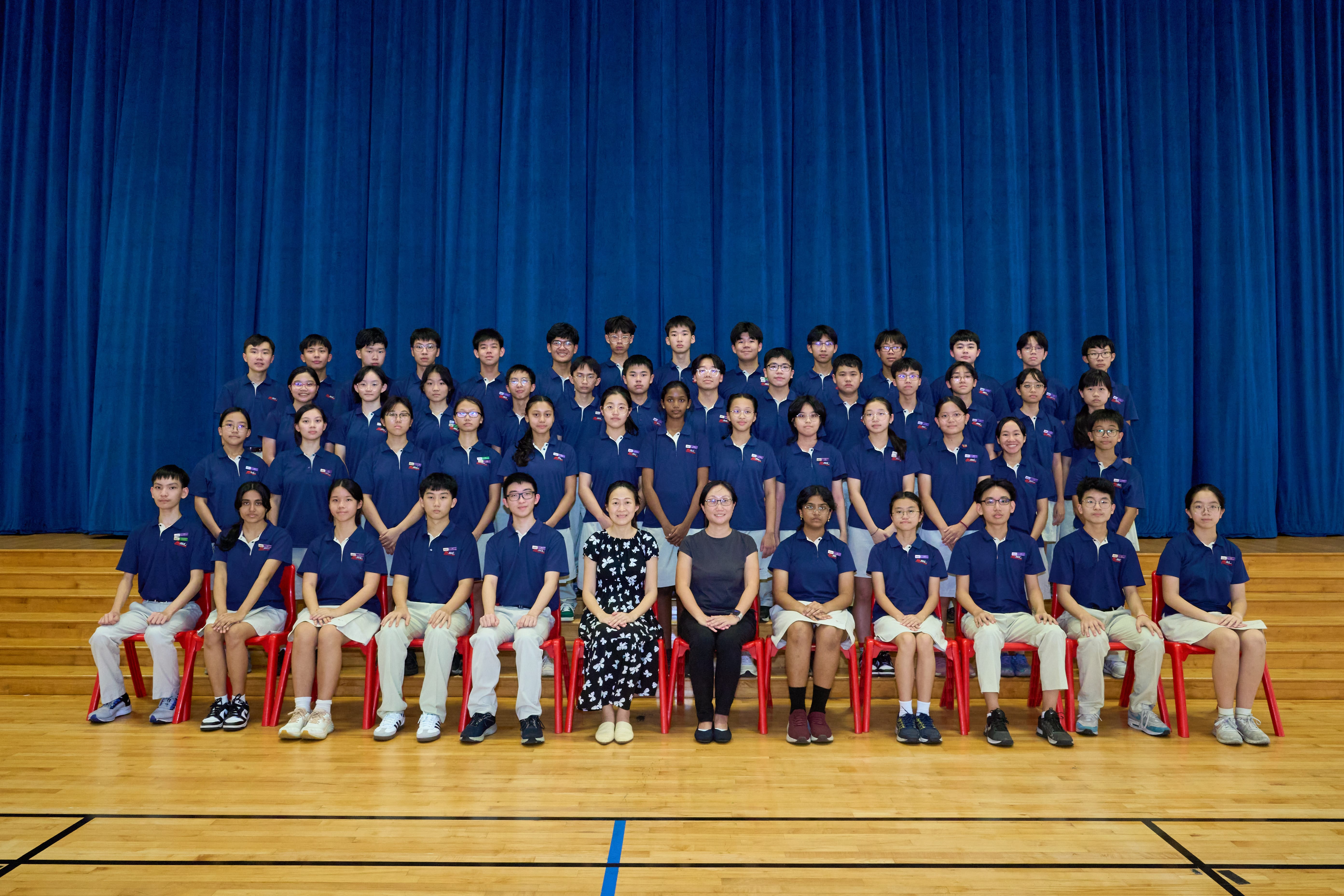 Group of students in navy uniforms posed in rows with two seated teachers, blue curtain backdrop.