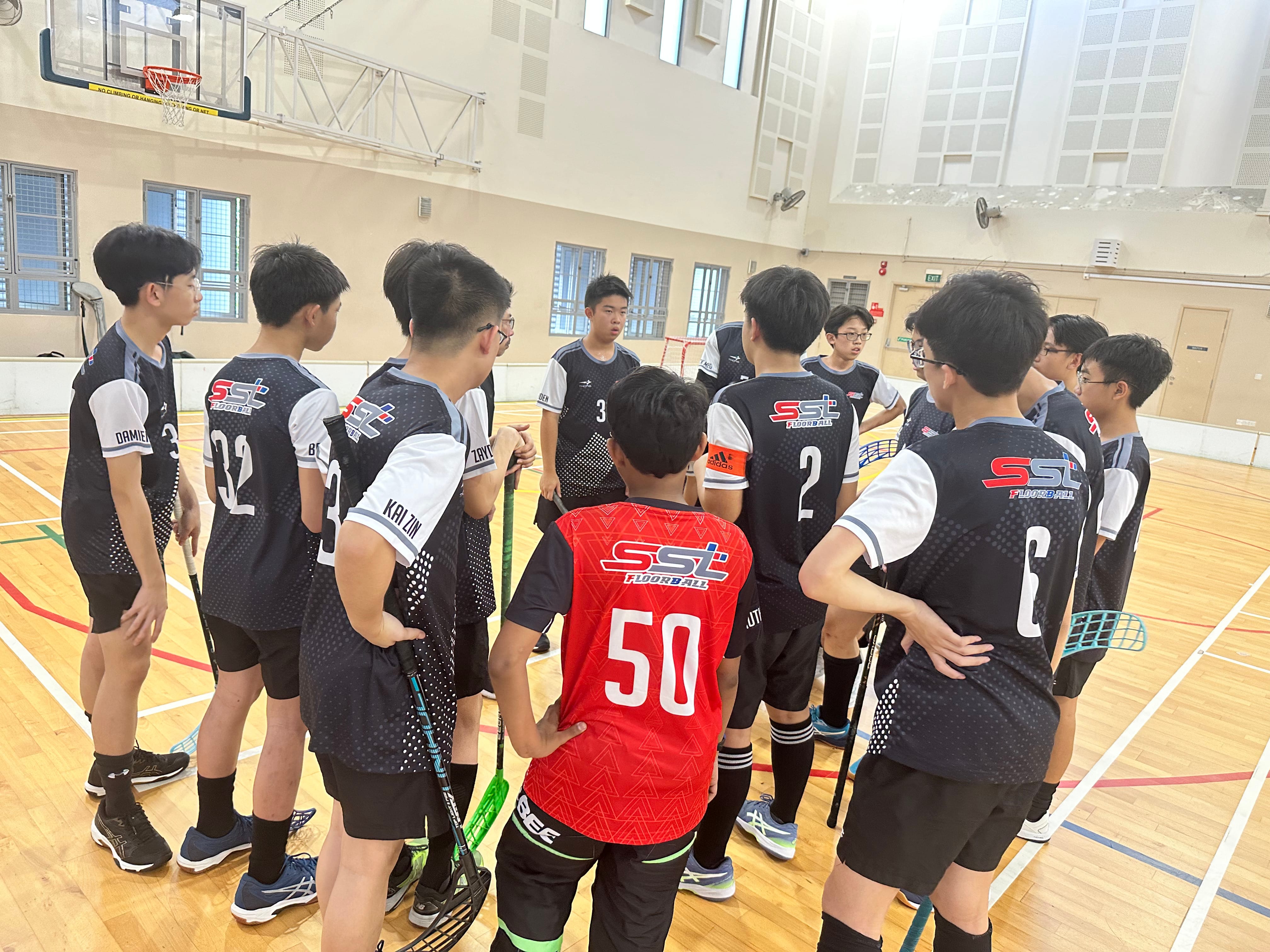 A youth floorball team in black SST jerseys huddles together on an indoor gymnasium court.