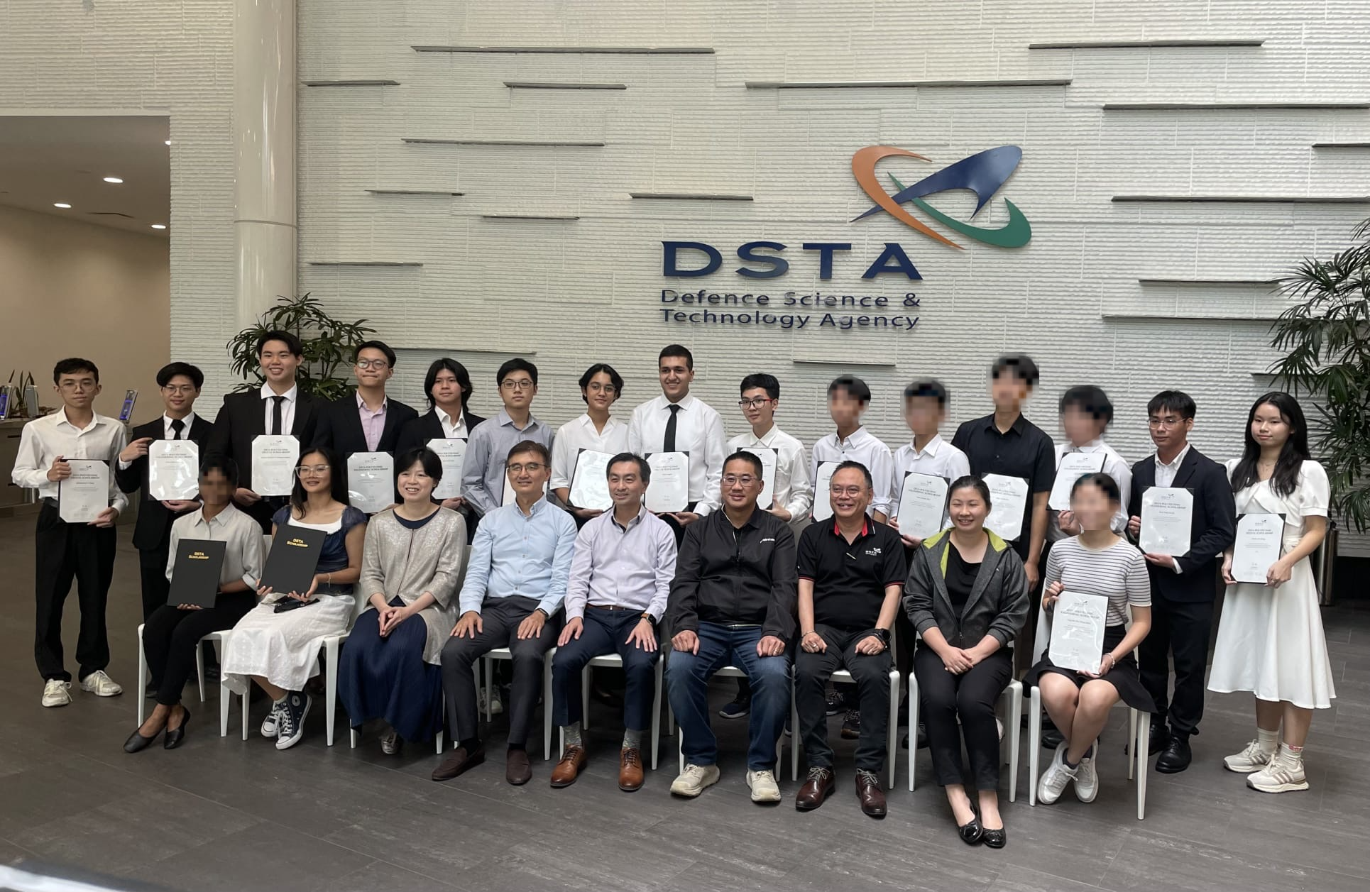 A group of people holding certificates pose in front of the DSTA Defence Science and Technology Agency sign.