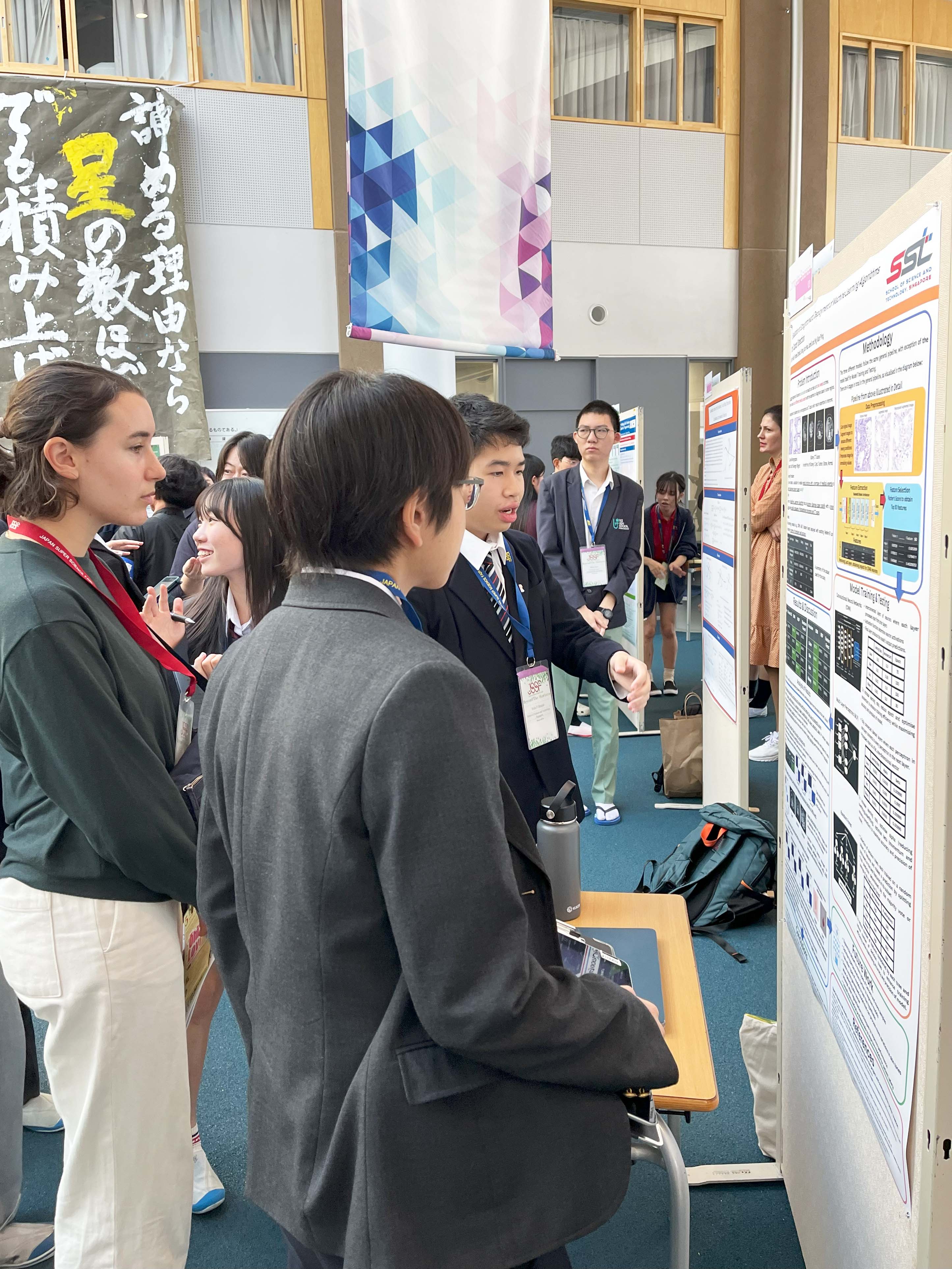 Students in uniforms and lanyards gather around a research poster board at an academic fair.