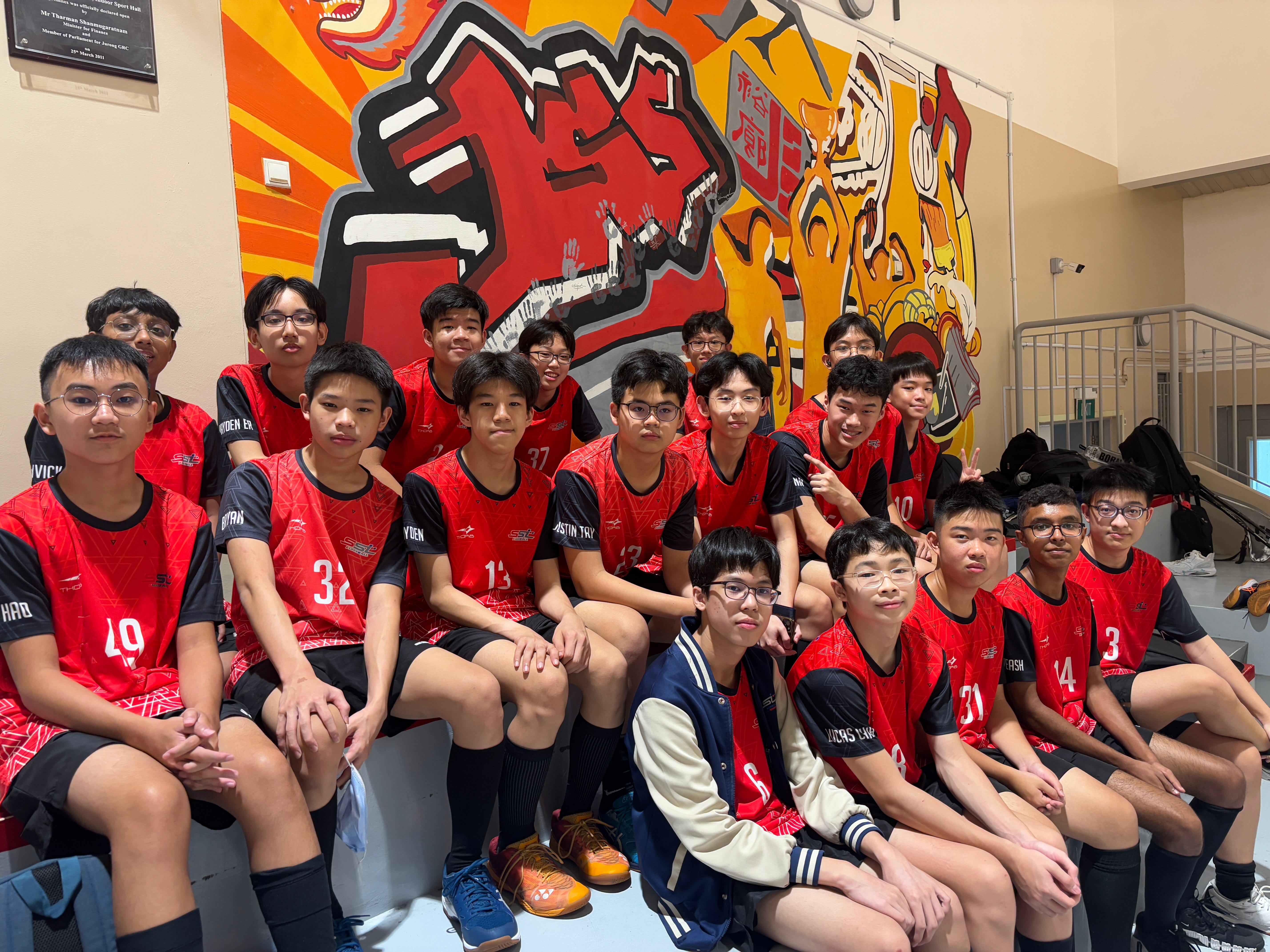 Group of teen boys in red sports jerseys seated on bleachers, with a bright mural behind them.