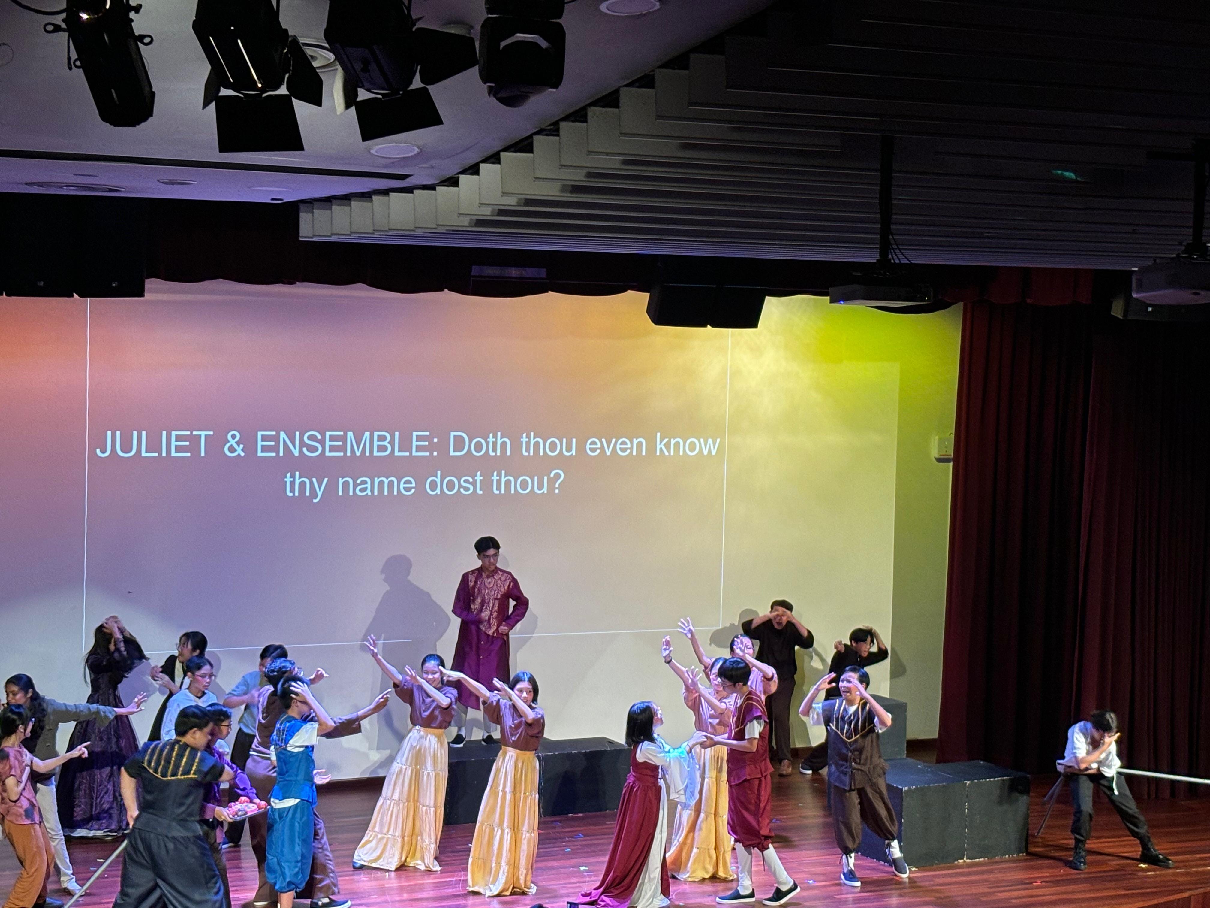 Students perform a Romeo and Juliet stage production with colorful costumes, projected lyrics visible on the backdrop.