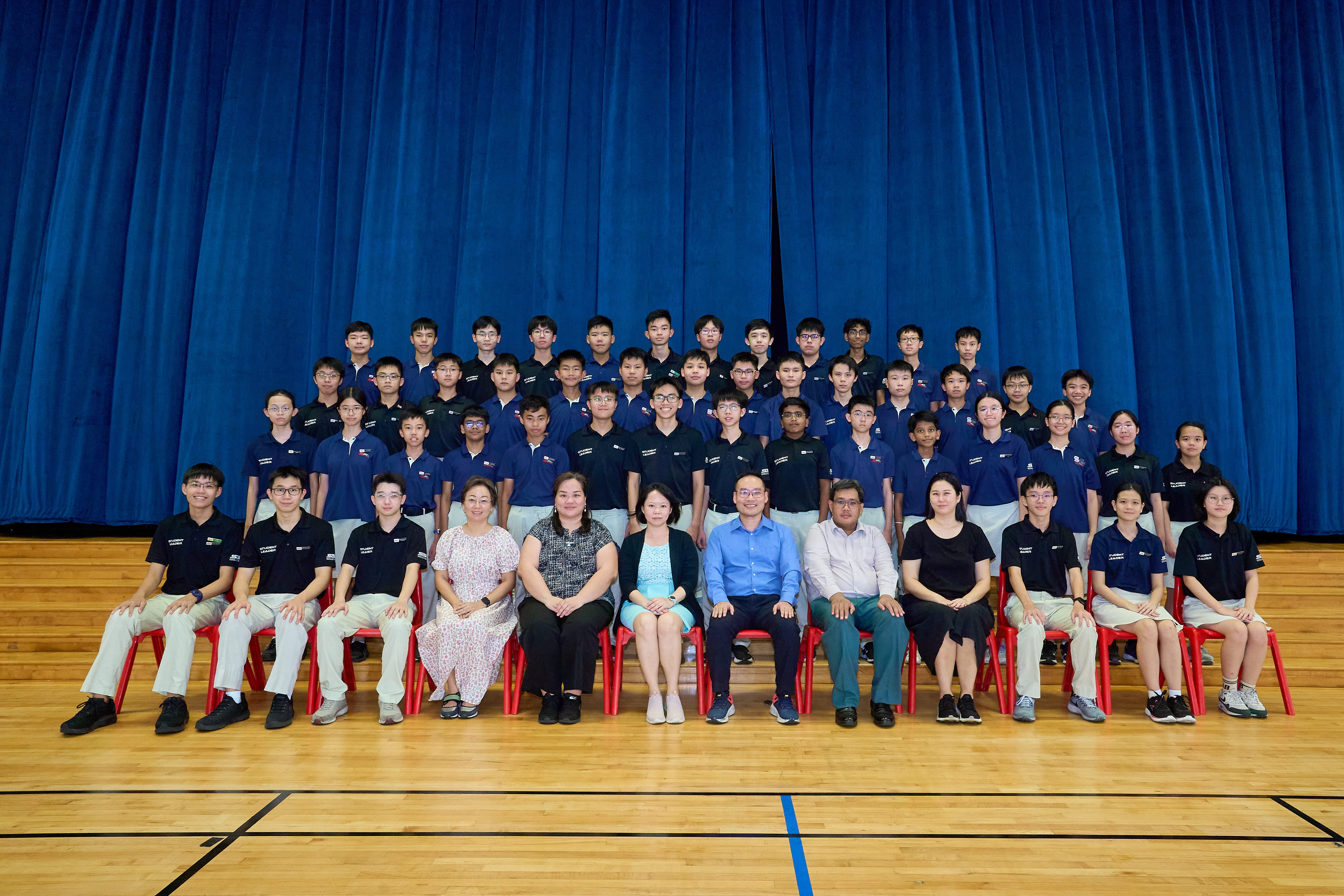 Large school group photo in a hall, students in navy uniforms with teachers seated in front of blue curtains.