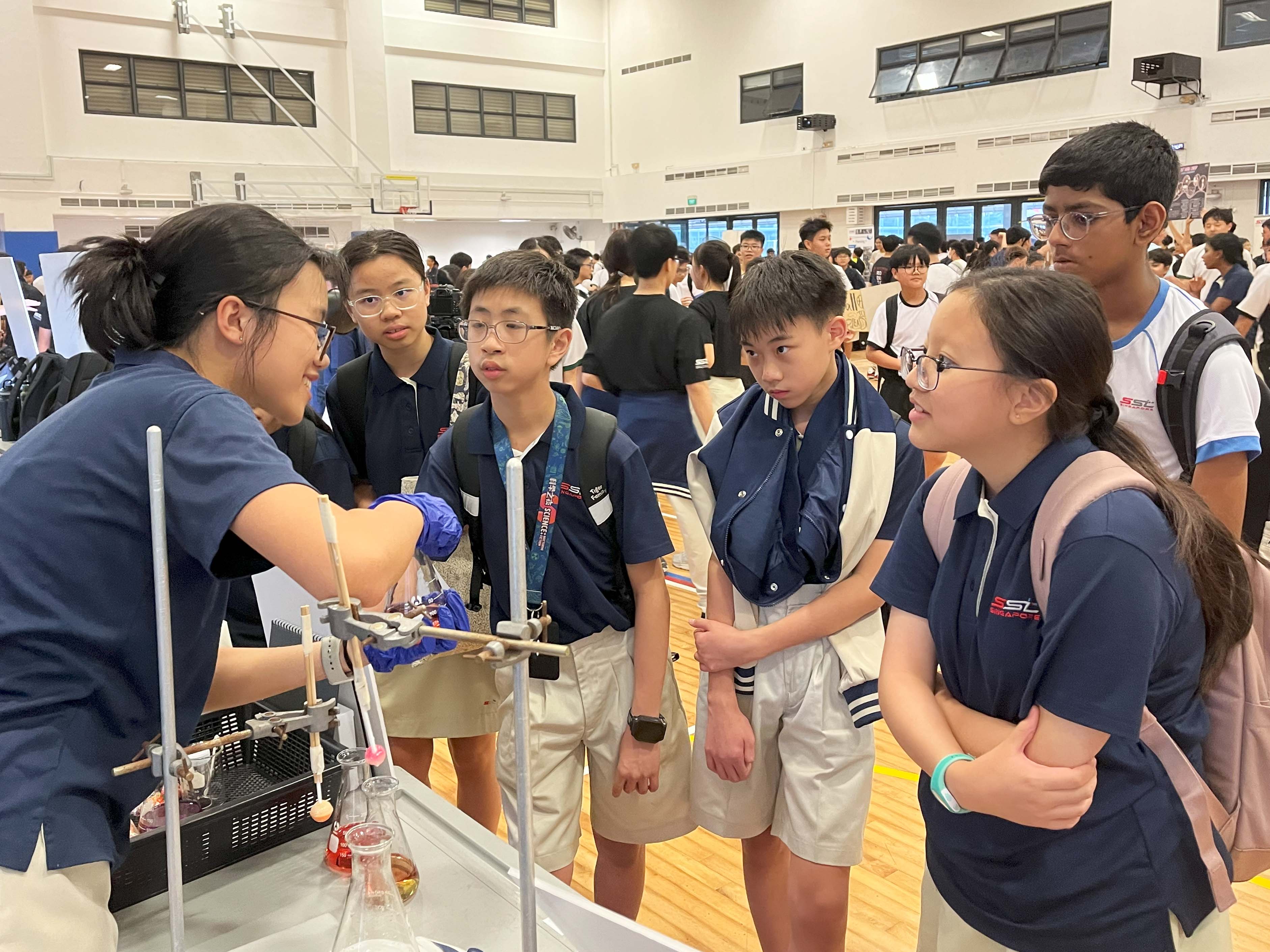 Students in navy uniforms watch a peer demonstrate a science experiment with lab equipment and flasks at a school fair.