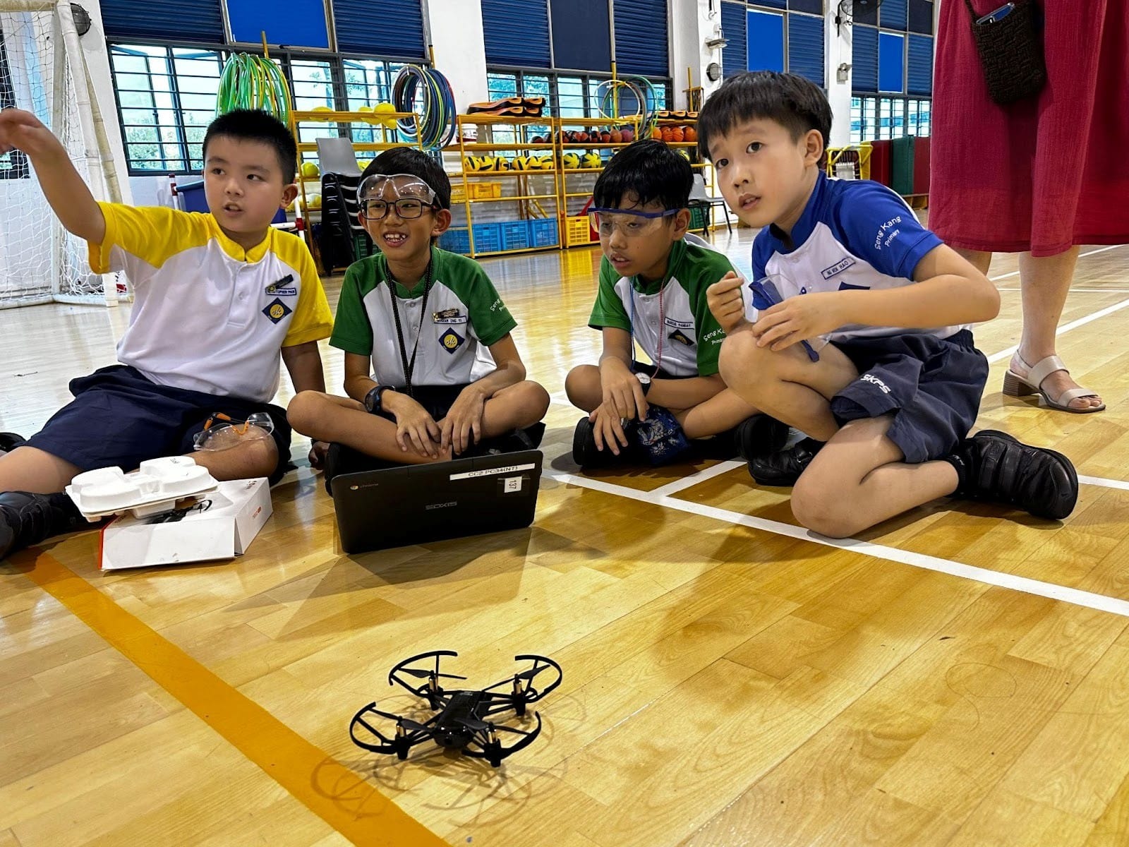 Four young boys in school uniforms sit on a gym floor with a small drone and a laptop in front of them.