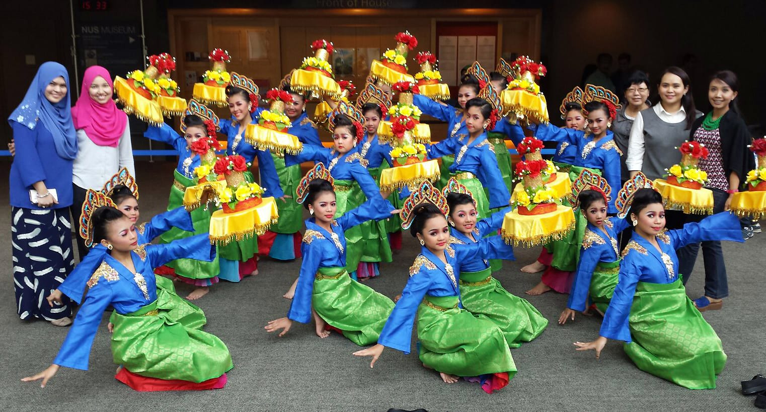 A large group of female dancers in blue and green costumes with tall floral yellow headdresses pose at NUS Museum.