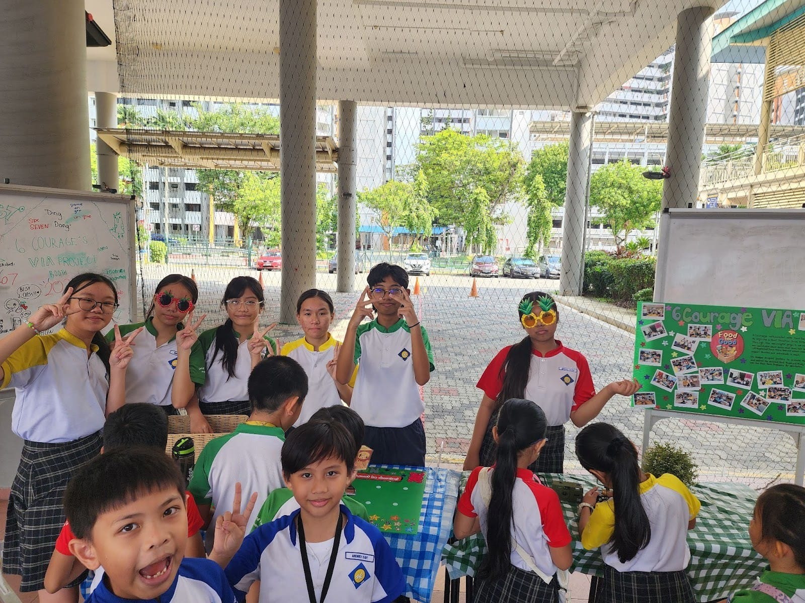 A group of school children in uniforms pose with peace signs and novelty glasses at an outdoor project booth.