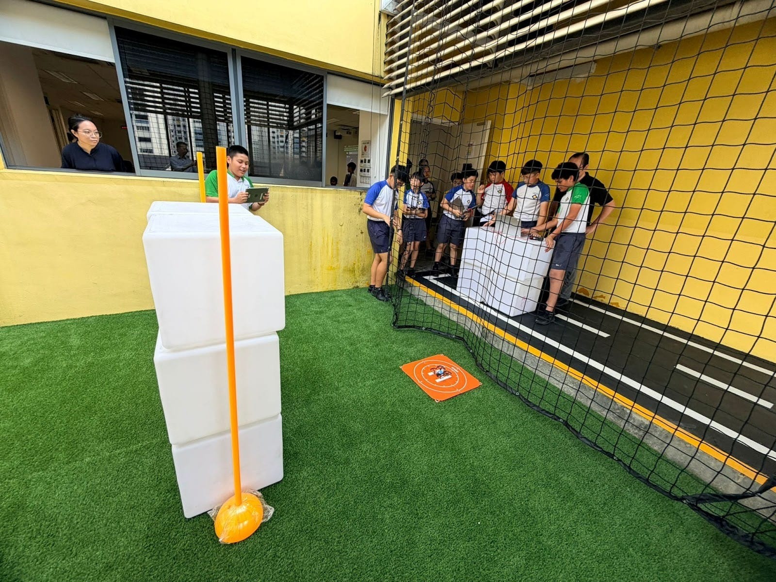 Students gather near a safety net on artificial turf beside white foam blocks, orange poles, and a small drone landing pad.