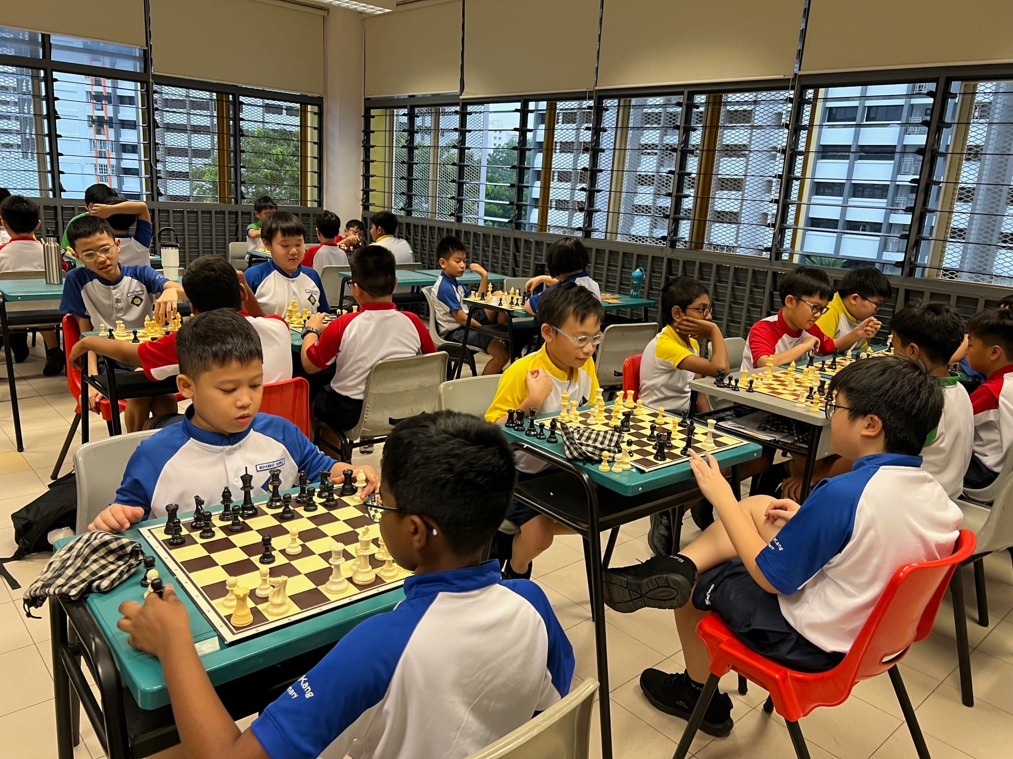 Many young boys in school uniforms sit at desks playing chess in a bright indoor room with large windows.