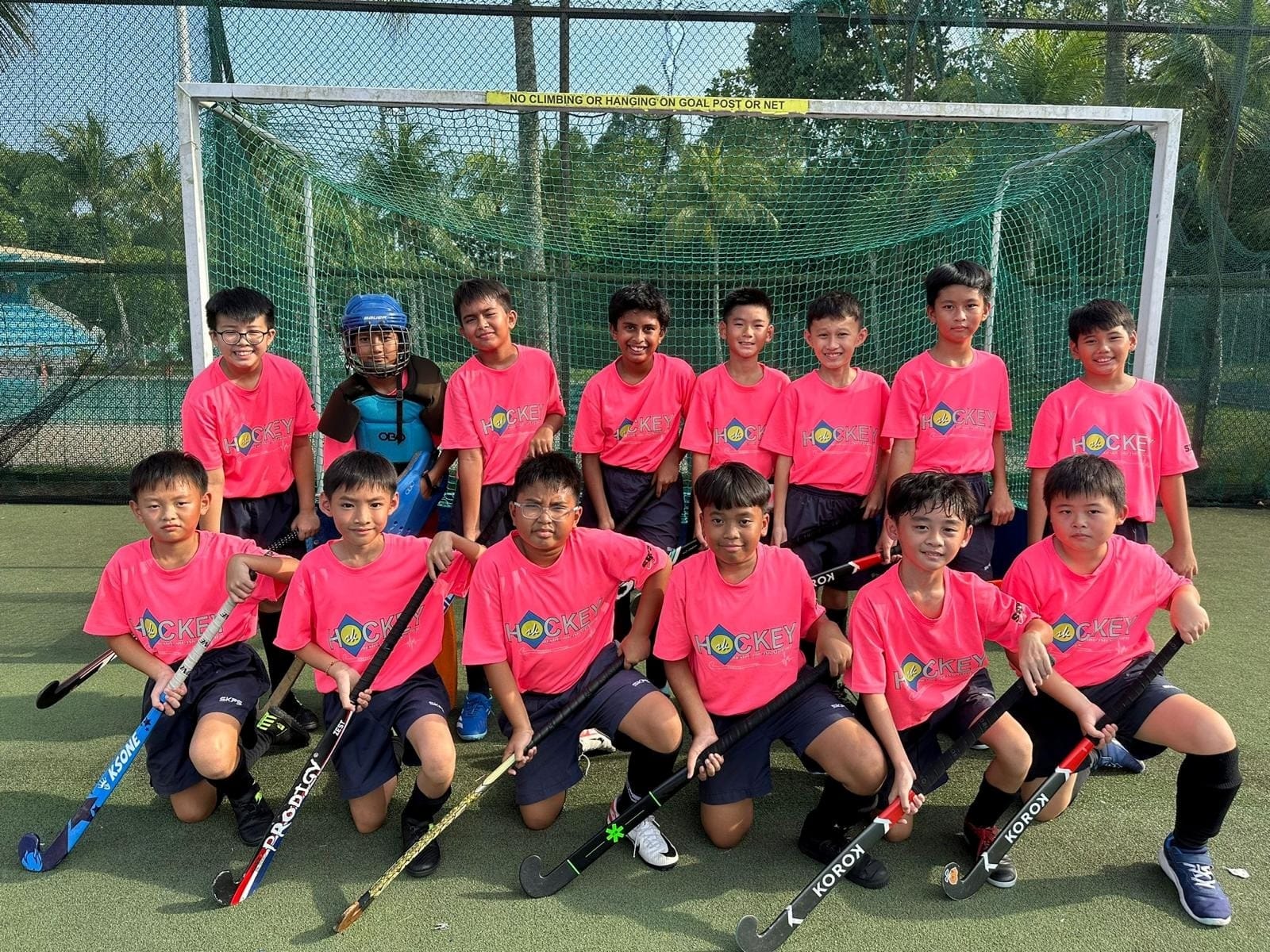 Young boys in pink hockey jerseys pose with sticks in front of a goal net, one wearing a blue goalkeeper helmet.