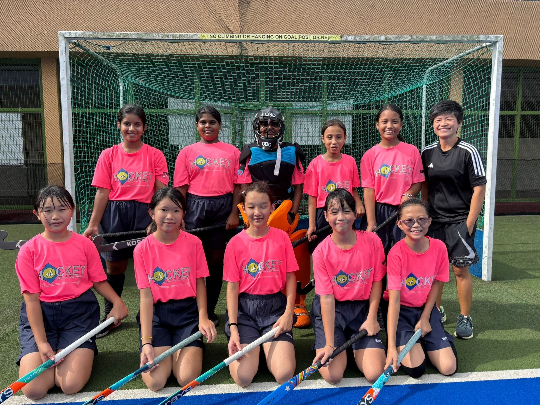 Girls field hockey team in pink jerseys poses in front of a goal, one player in goalie gear, coach in black.