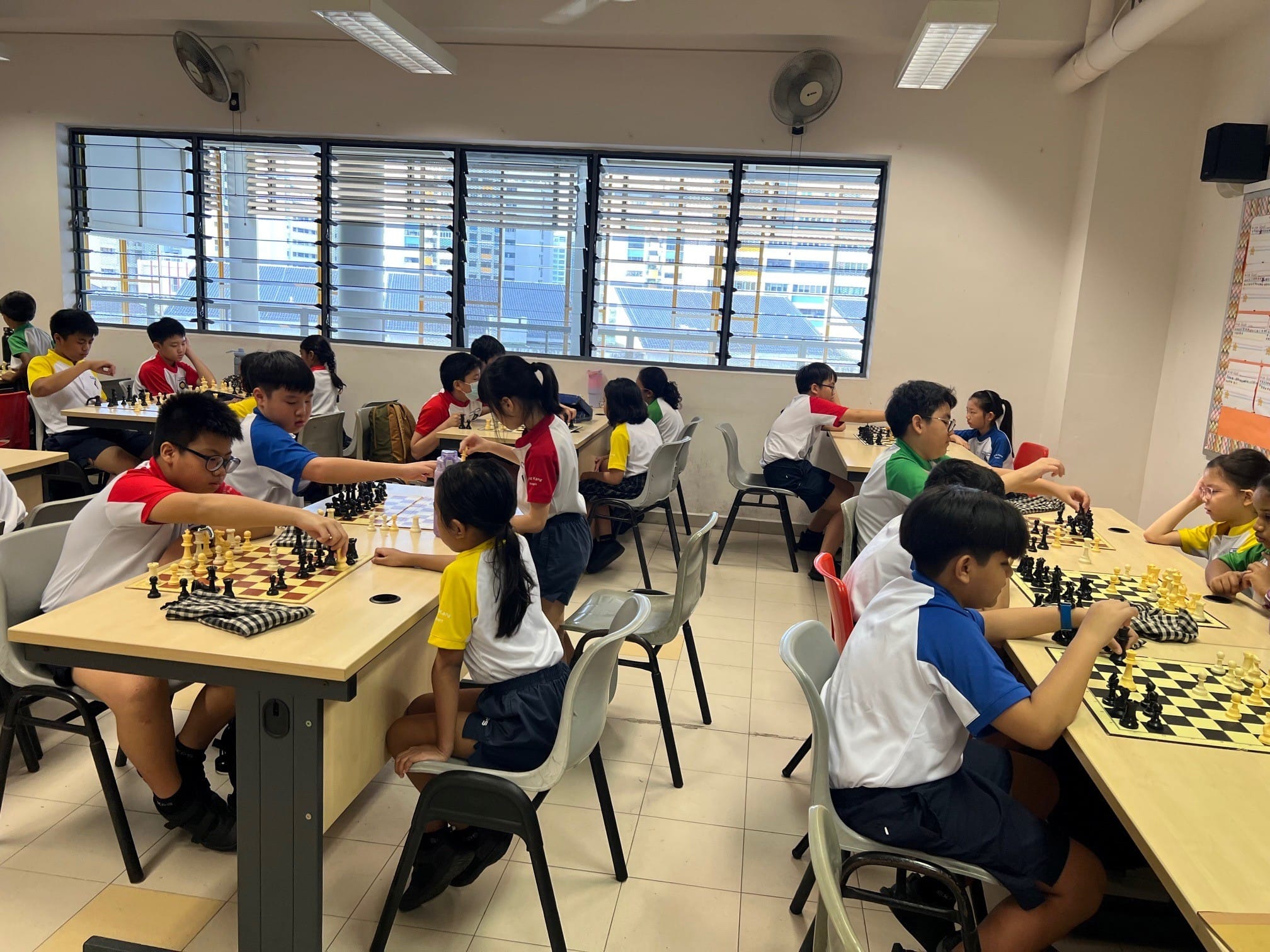Children in school uniforms play chess at tables in a bright classroom with louvered windows.