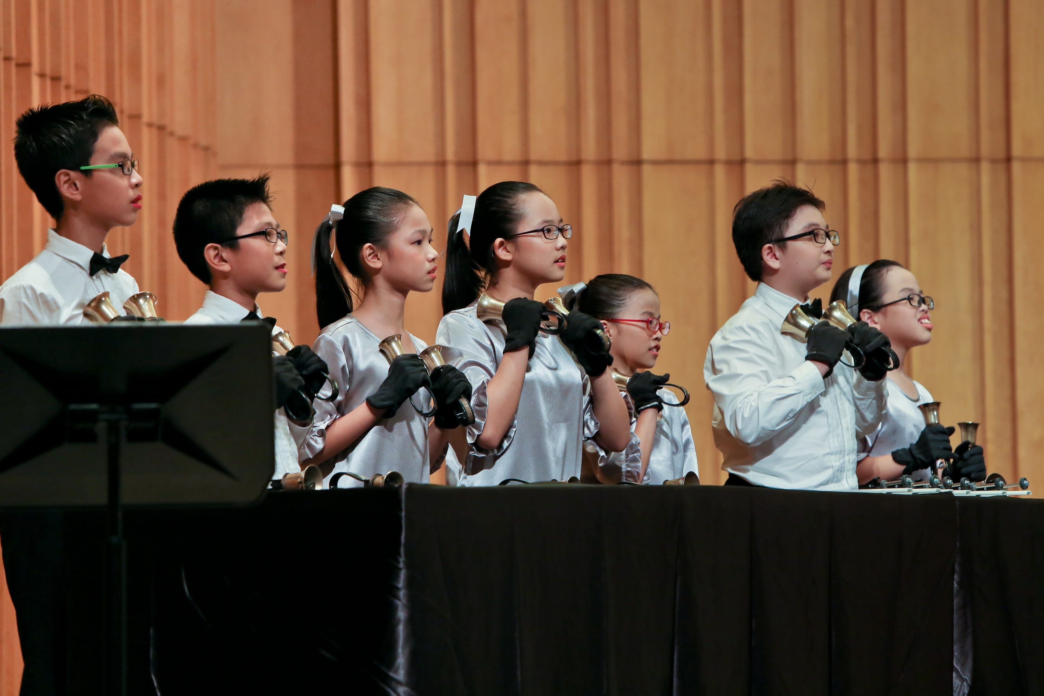 Seven children in white outfits wearing black gloves hold handbells during a stage performance.
