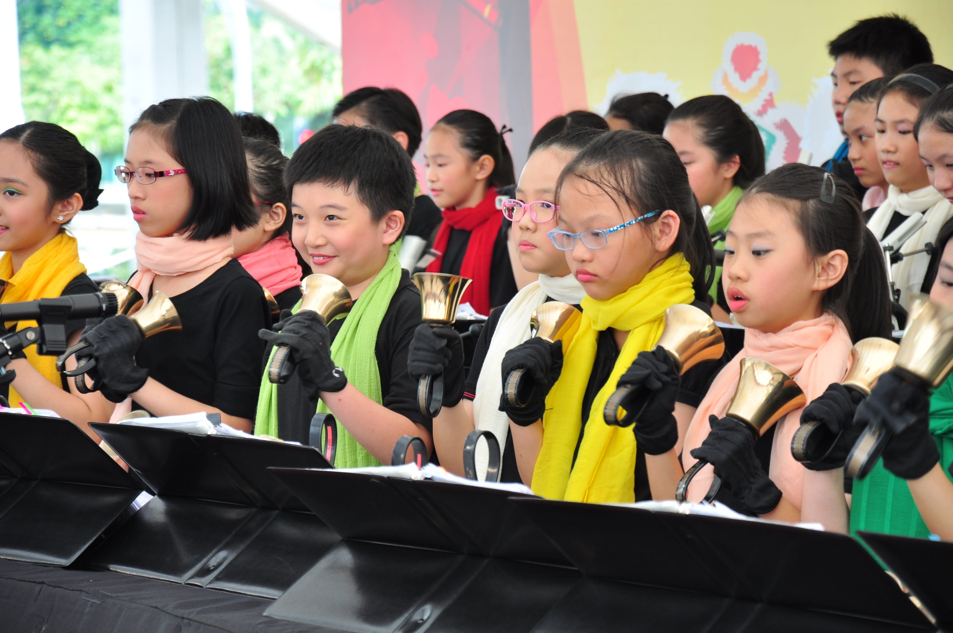 Children wearing black gloves and colorful scarves hold gold handbells during an outdoor performance.