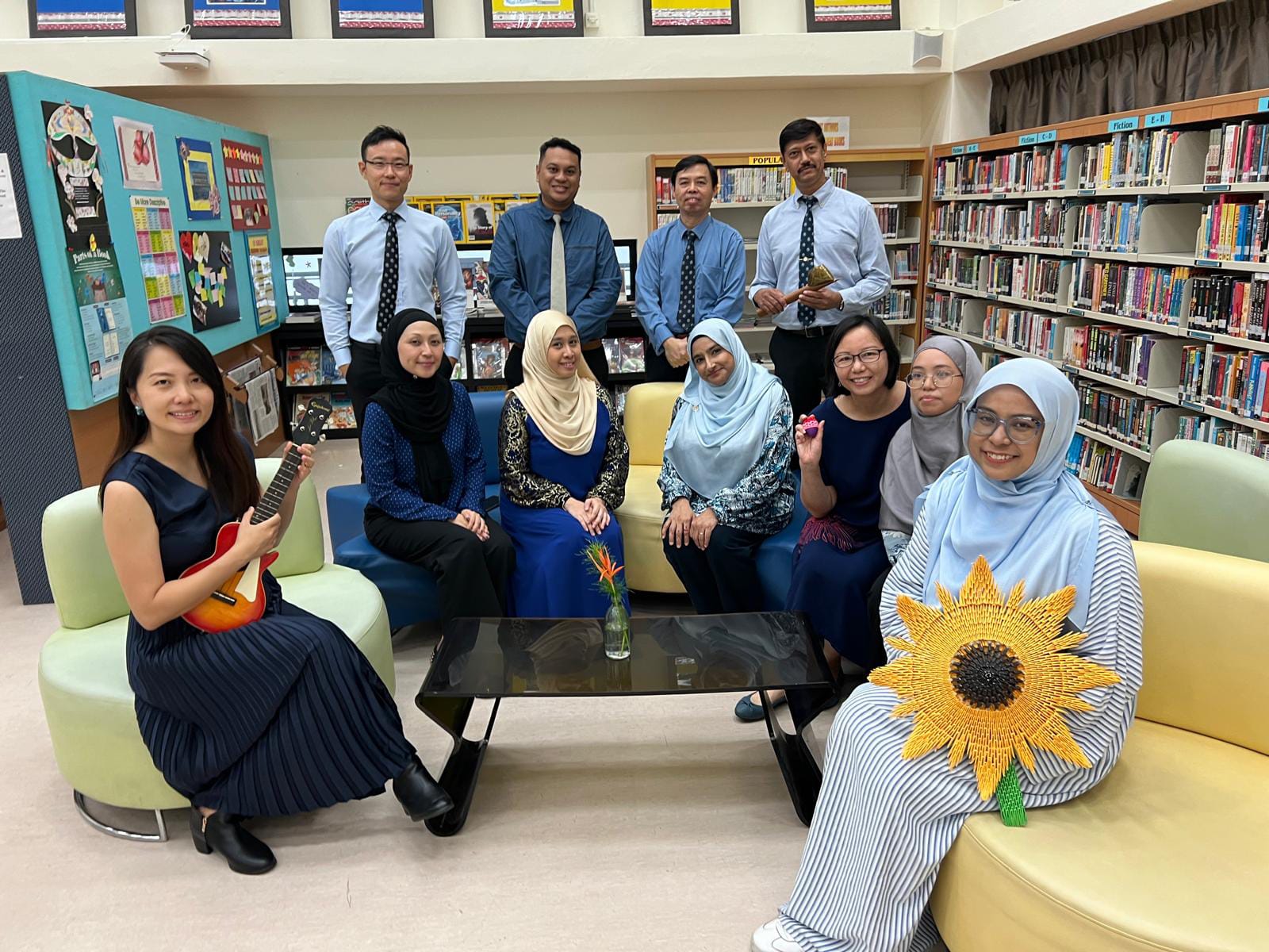 Ten adults pose in a school library; one holds a ukulele, another holds a large sunflower decoration.
