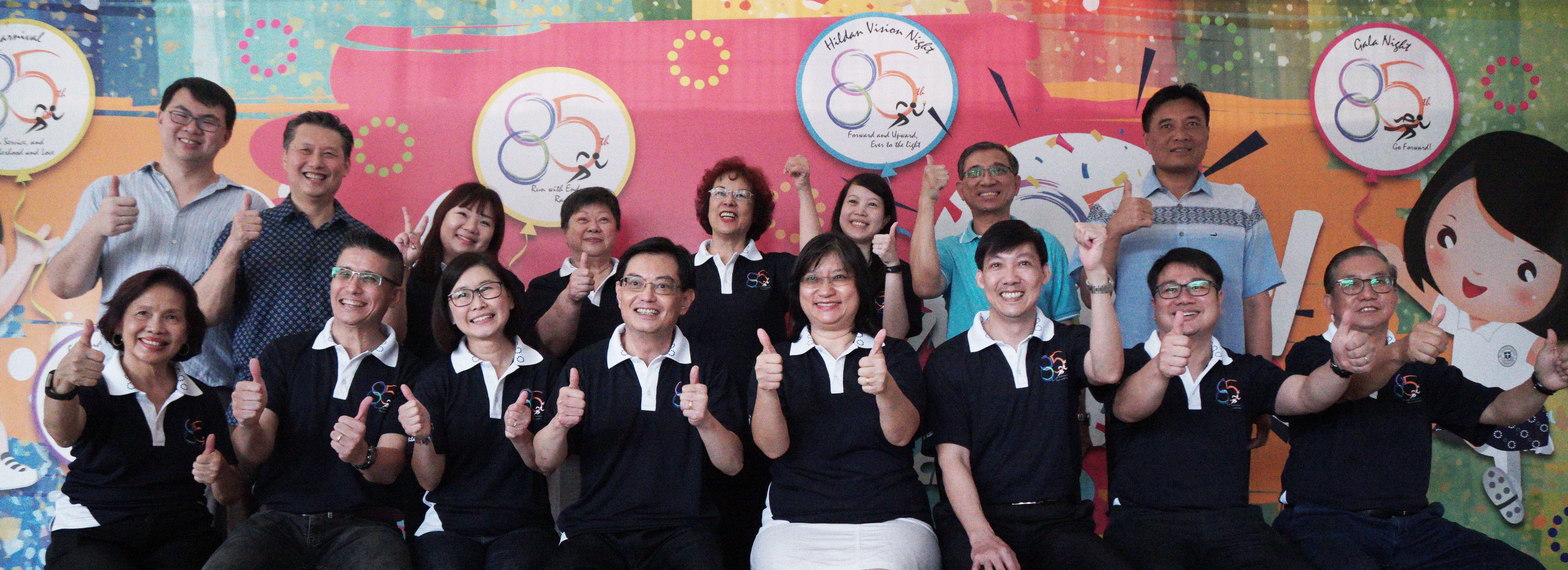 Group of 14 smiling adults giving thumbs-up in front of a colorful 85th anniversary backdrop.