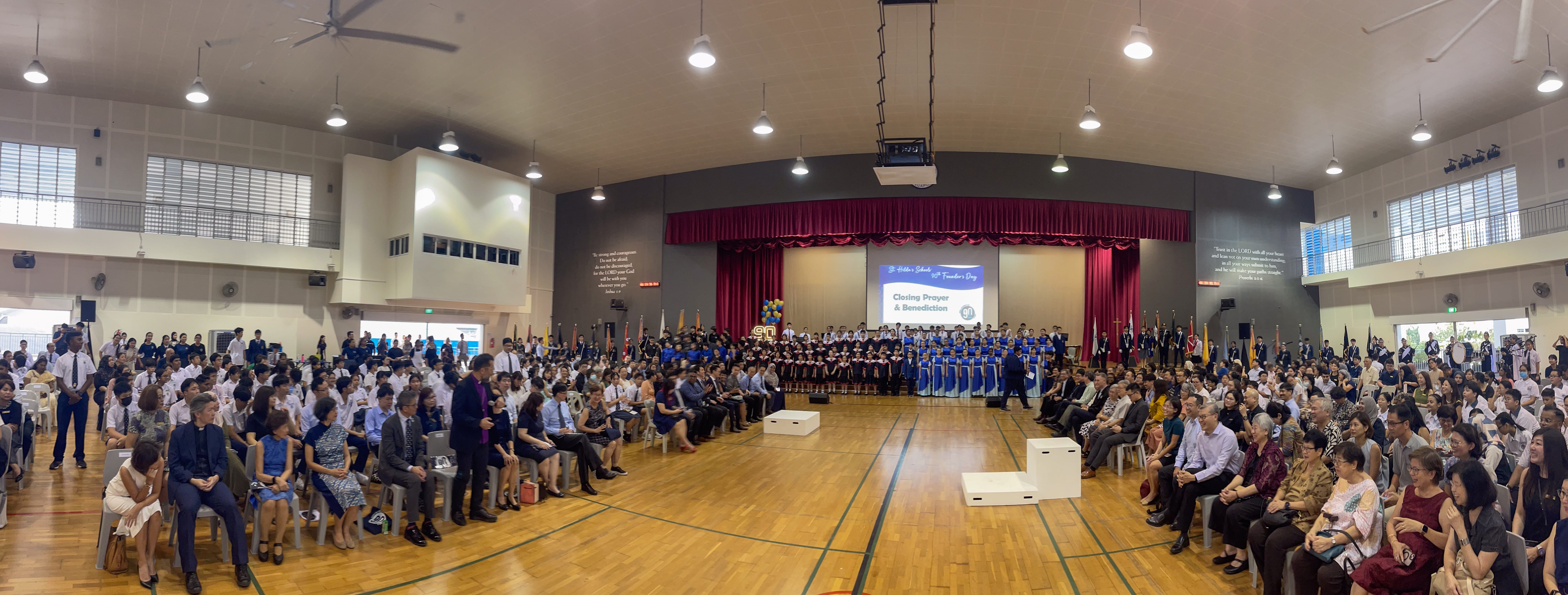 Large indoor auditorium with seated audience facing a stage with students in uniform and a closing prayer screen.