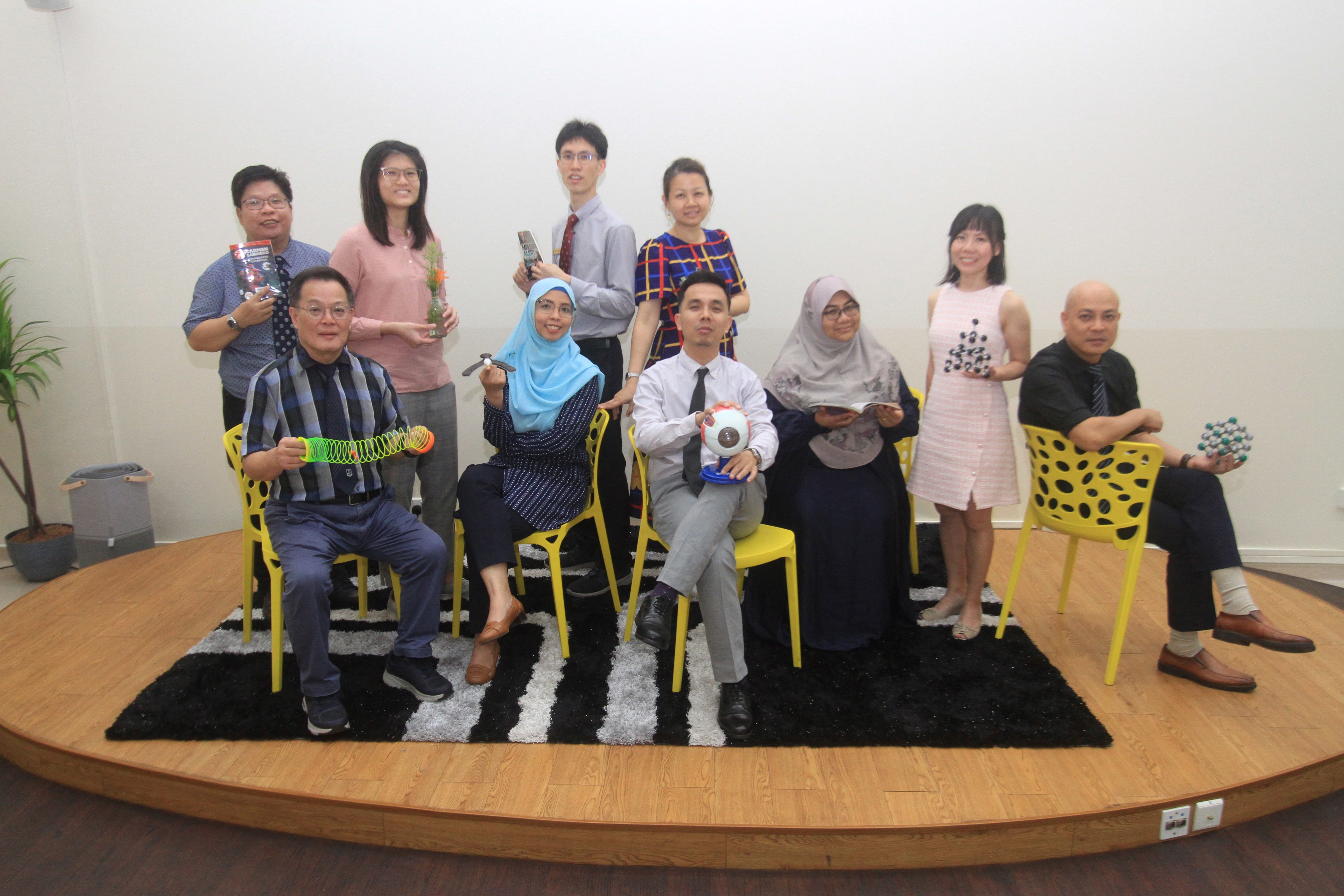 Ten people pose on a raised wooden platform with yellow chairs, holding science models and props like a slinky and eye model.