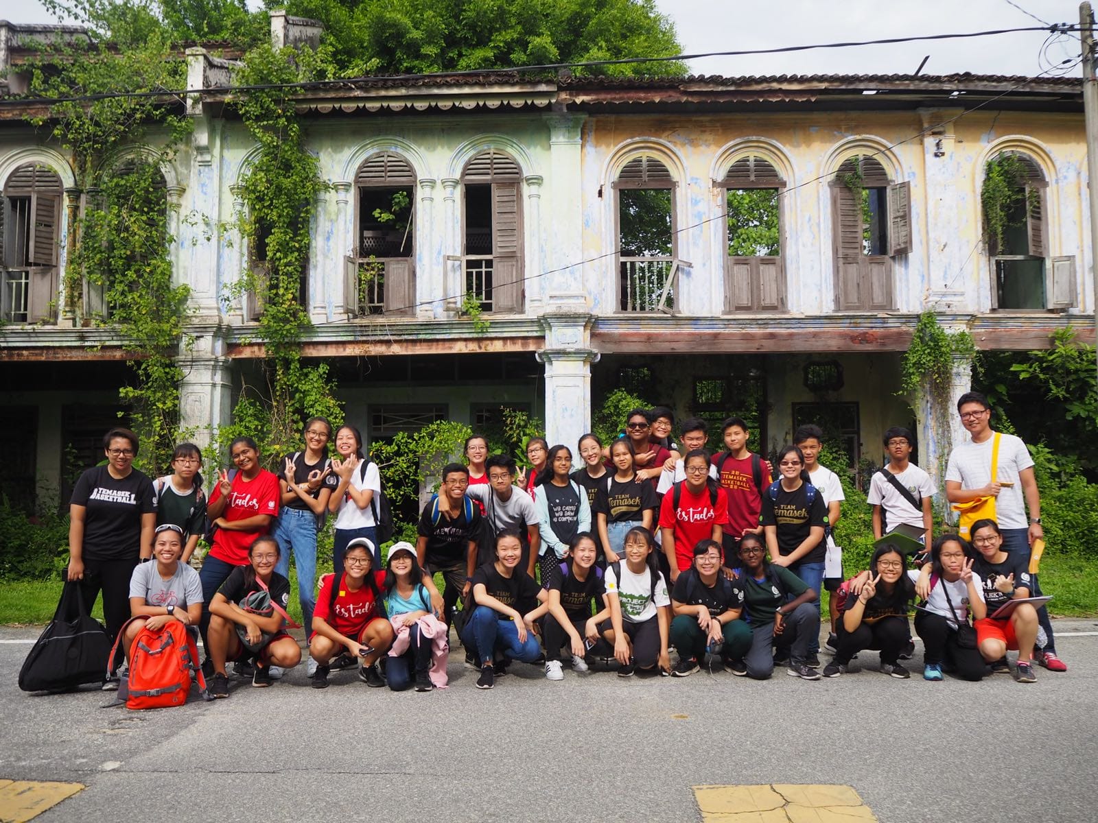 A large group of young people posing in front of a weathered, ivy-covered two-story colonial building.