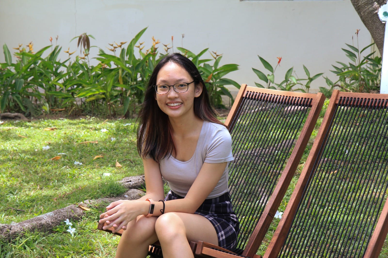 Young woman with glasses and long dark hair smiling, seated on a wooden chair outdoors with green plants behind her.