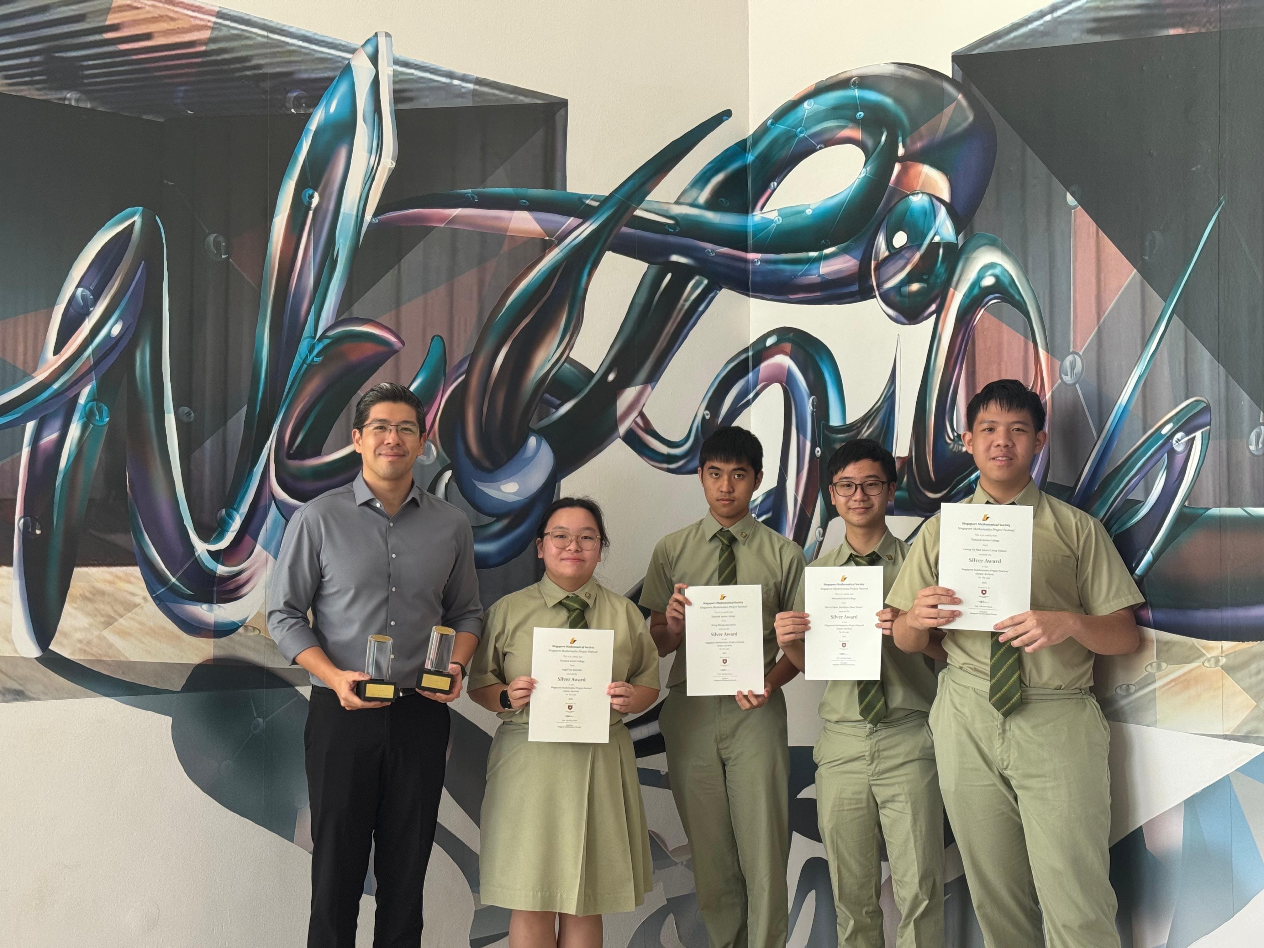 Five people pose before a colorful mural, four students in uniforms holding Silver Award certificates and one adult holding a trophy.