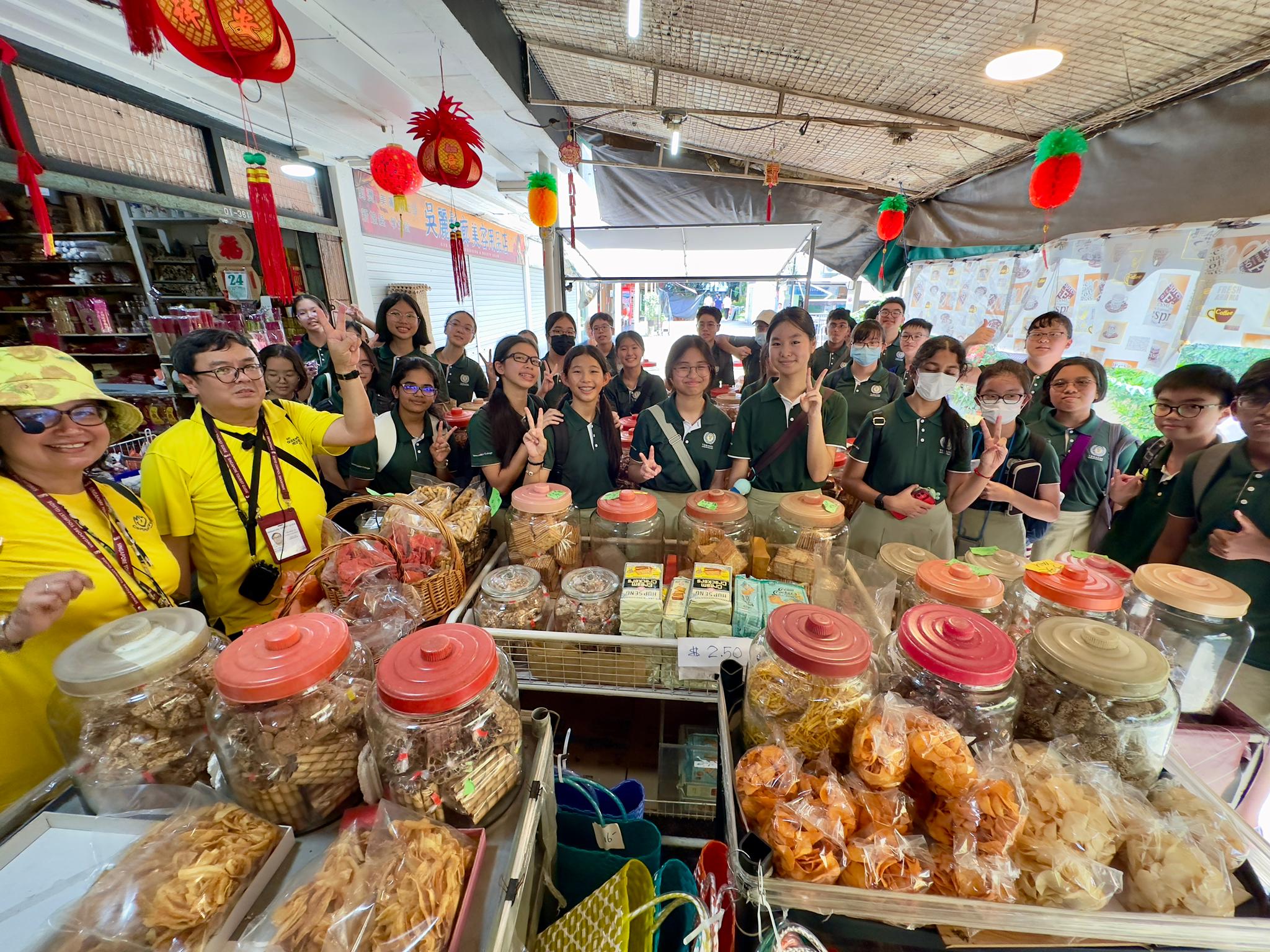 Students in green uniforms pose behind a market stall filled with snack jars and packaged foods, decorated with red Chinese lanterns.
