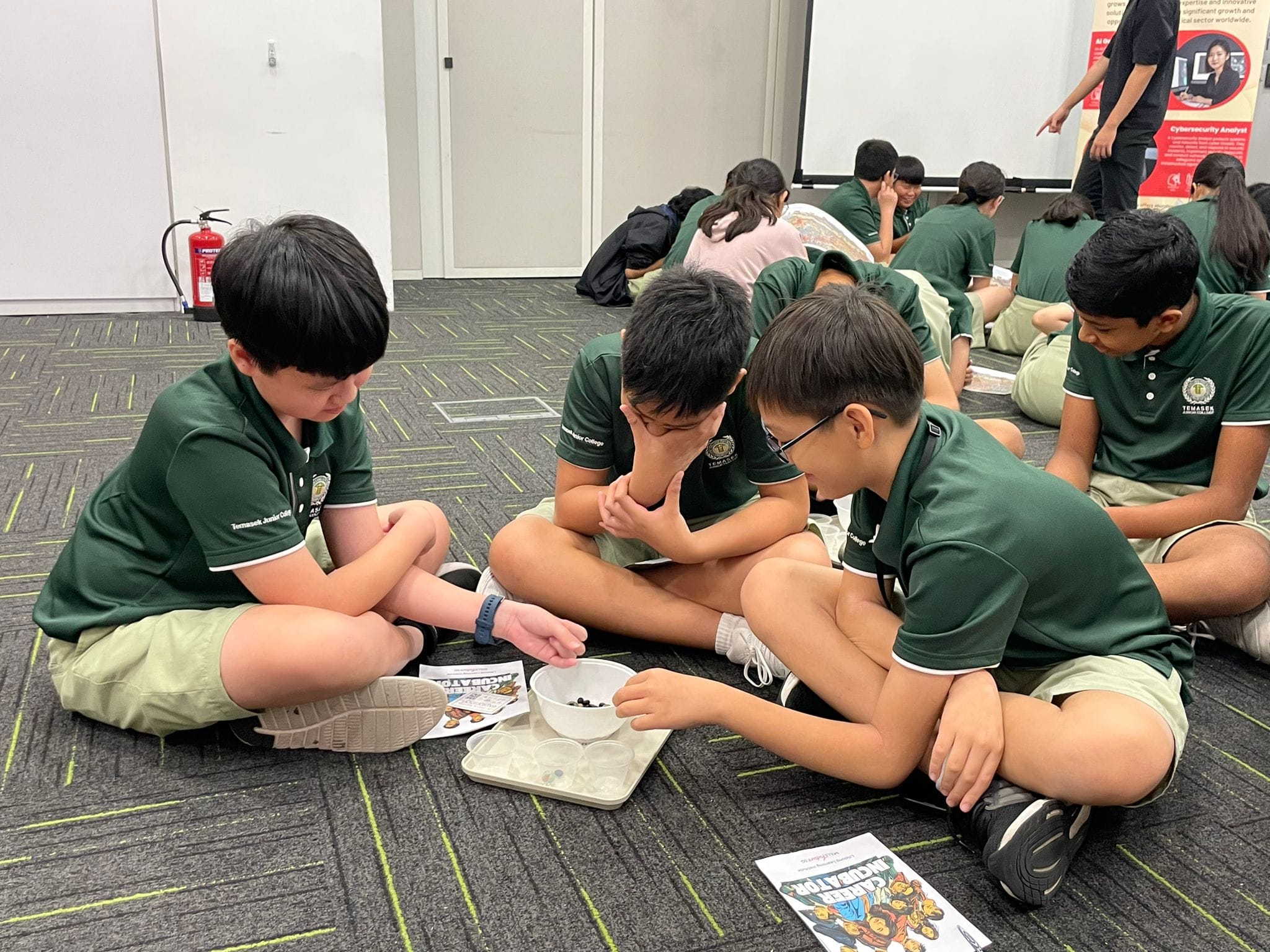 Students in green uniforms sit on a carpeted floor, examining small objects in a bowl during a group activity.