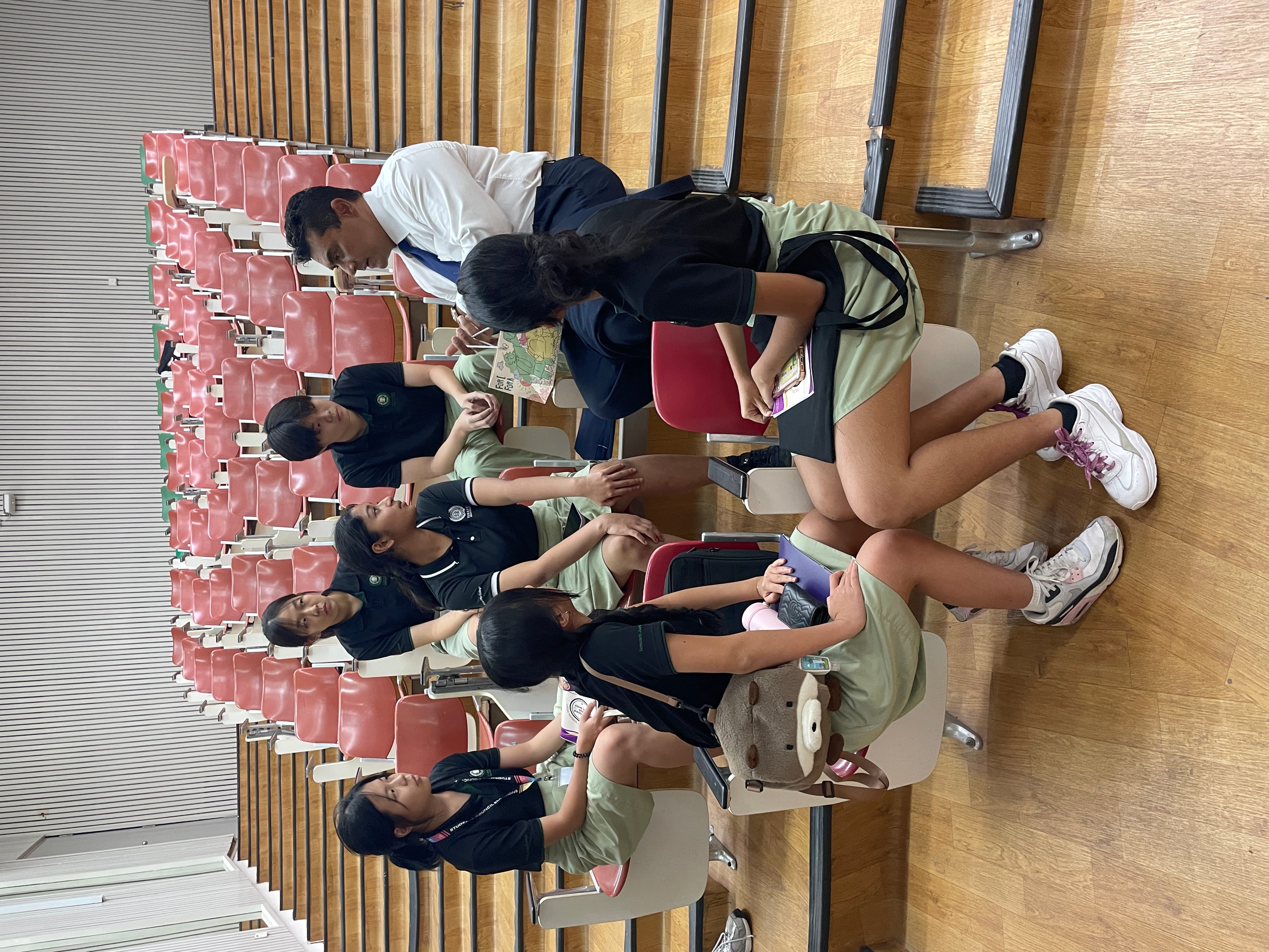 Students in uniforms sit in auditorium seats as a man leans in, showing them a book or paper.