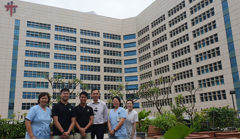 Six people stand smiling in front of a large multi-story hospital building with many windows.