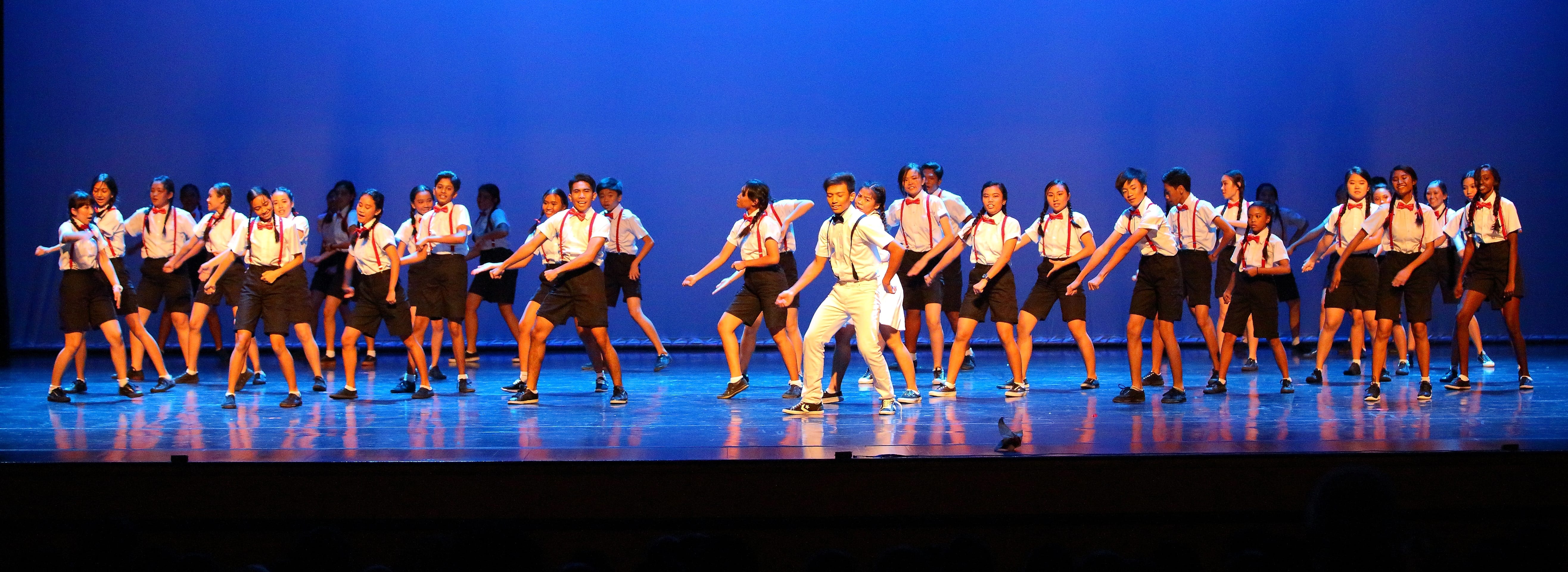 Large group of students in white shirts and suspenders dancing on a stage with blue lighting.