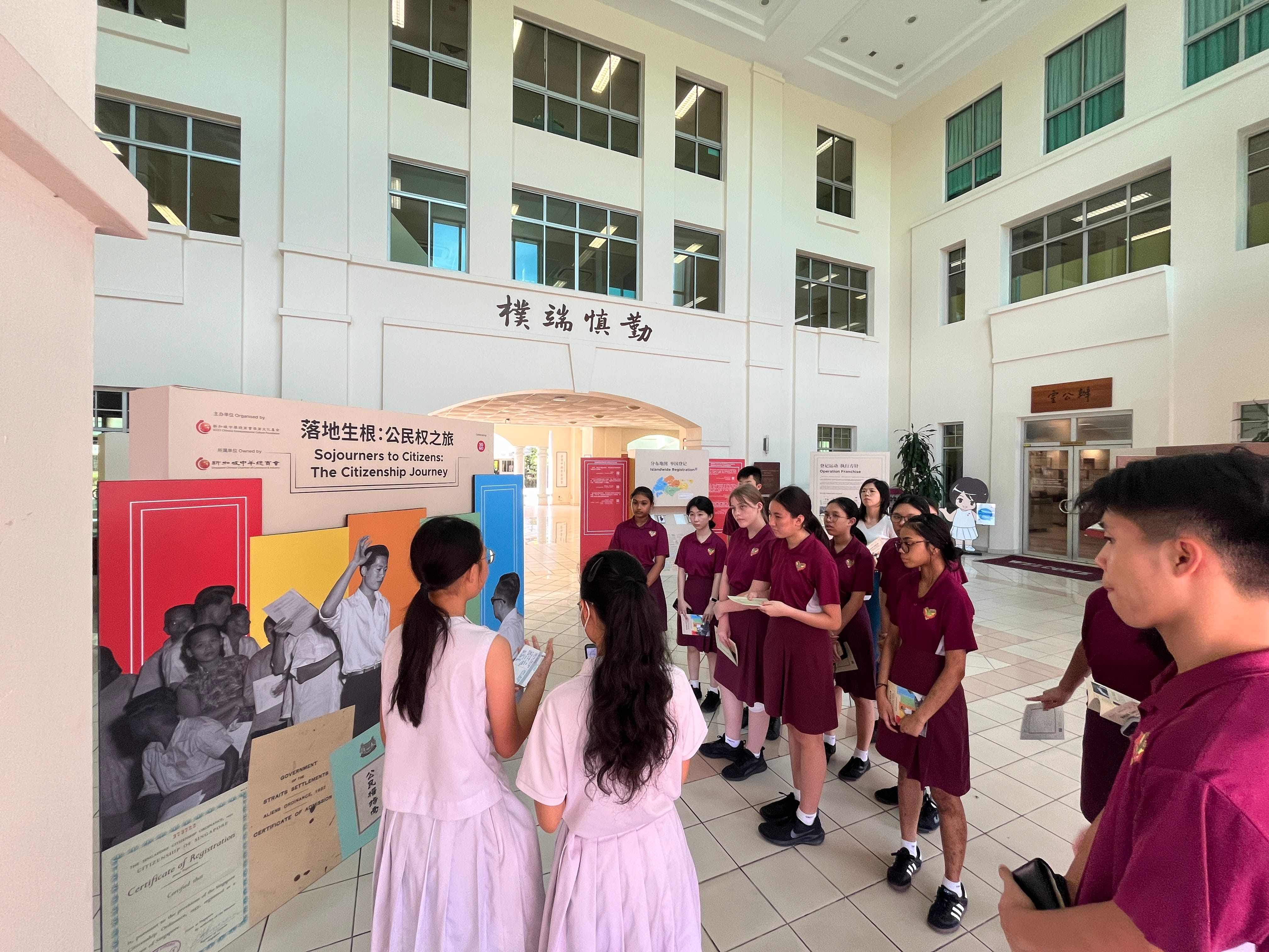 Students in uniforms view a colorful exhibition titled "Sojourners to Citizens: The Citizenship Journey" inside a hall.