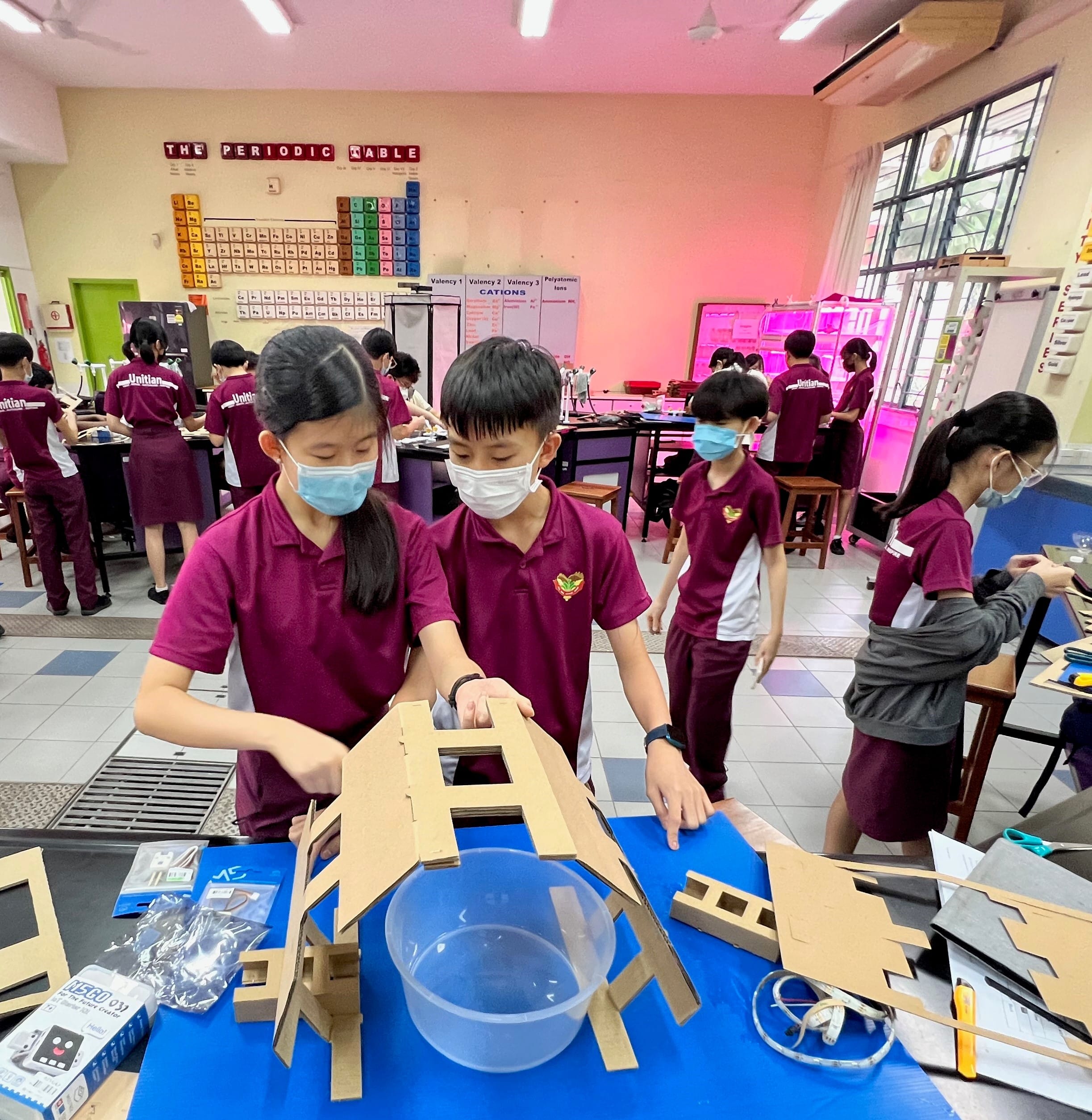 Students in maroon uniforms and masks assemble cardboard structures in a science classroom with a periodic table on the wall.