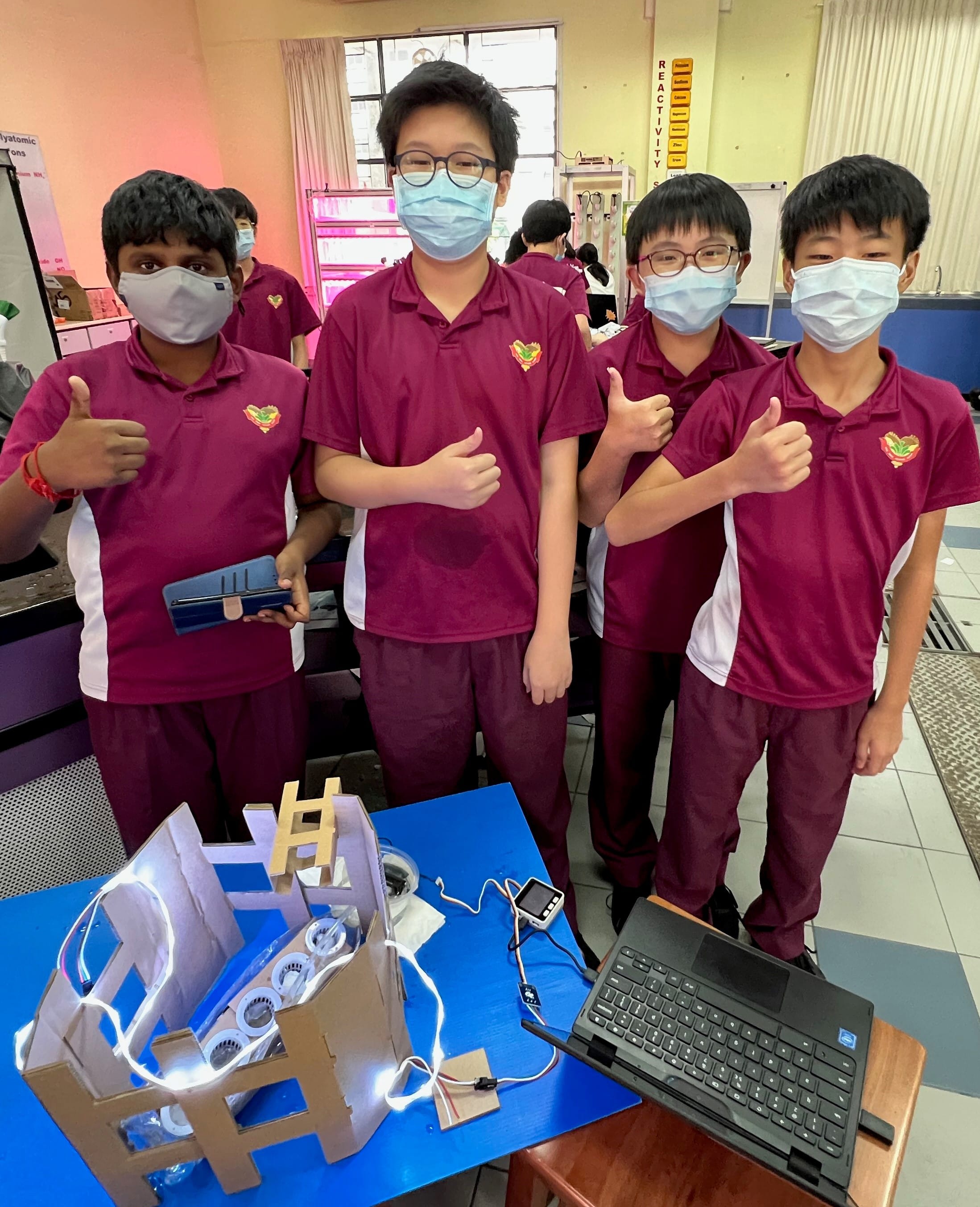 Four masked students in maroon uniforms give thumbs up beside a cardboard project with lights and a laptop.