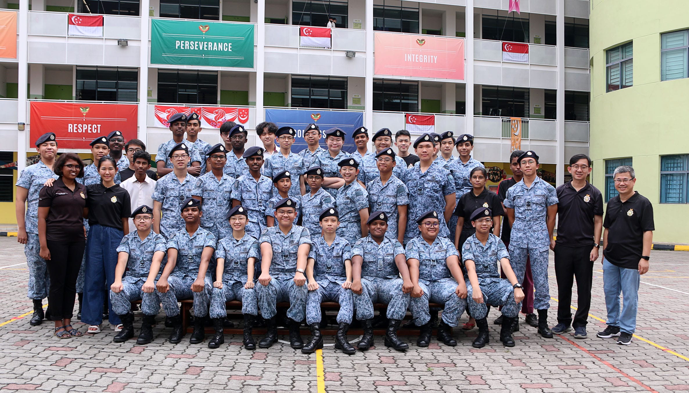 A large group of students in blue camouflage uniforms and berets pose outdoors in front of a school building.