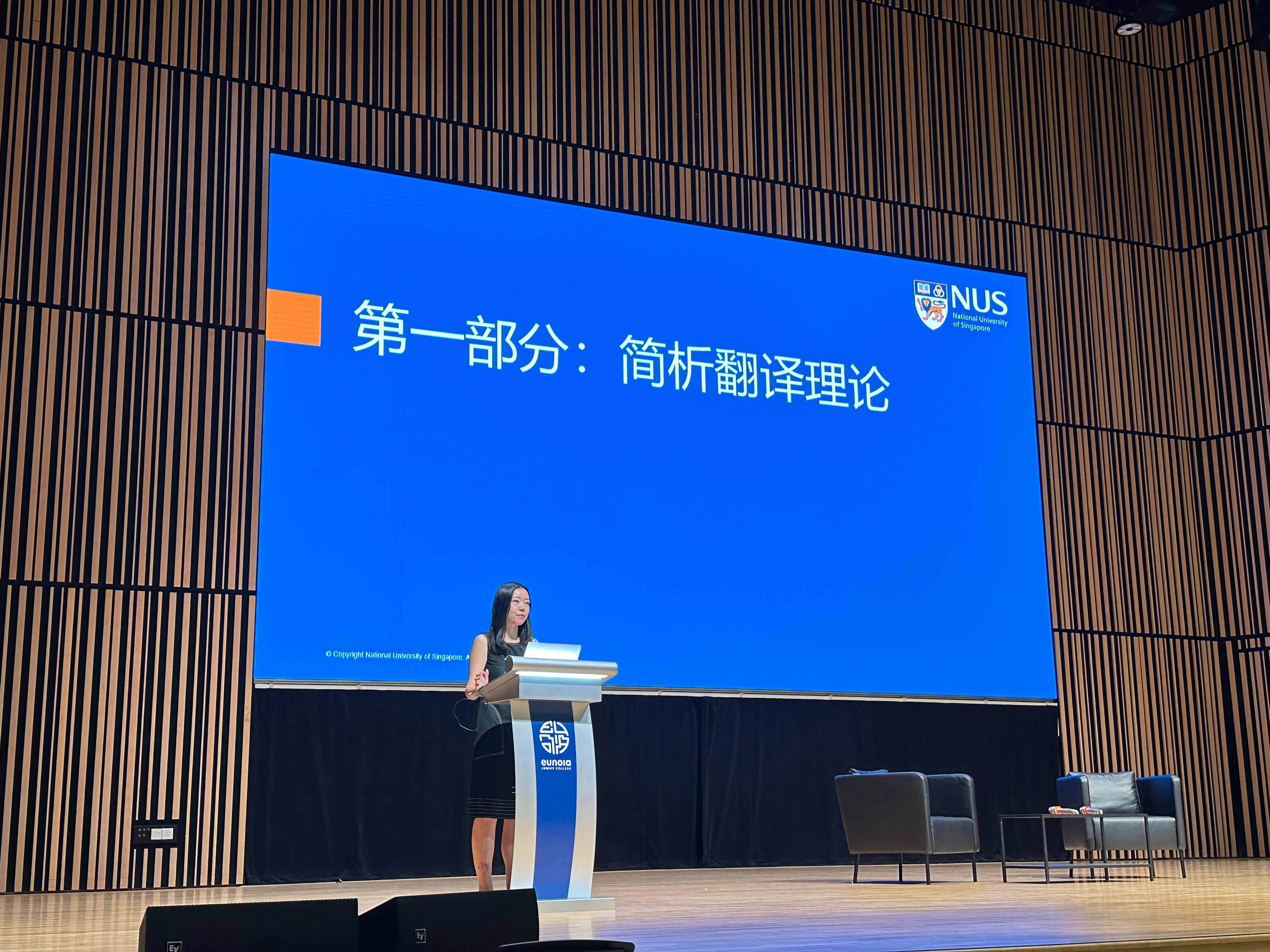 Woman speaking at a podium on stage before a blue NUS slide with Chinese text about translation theory.