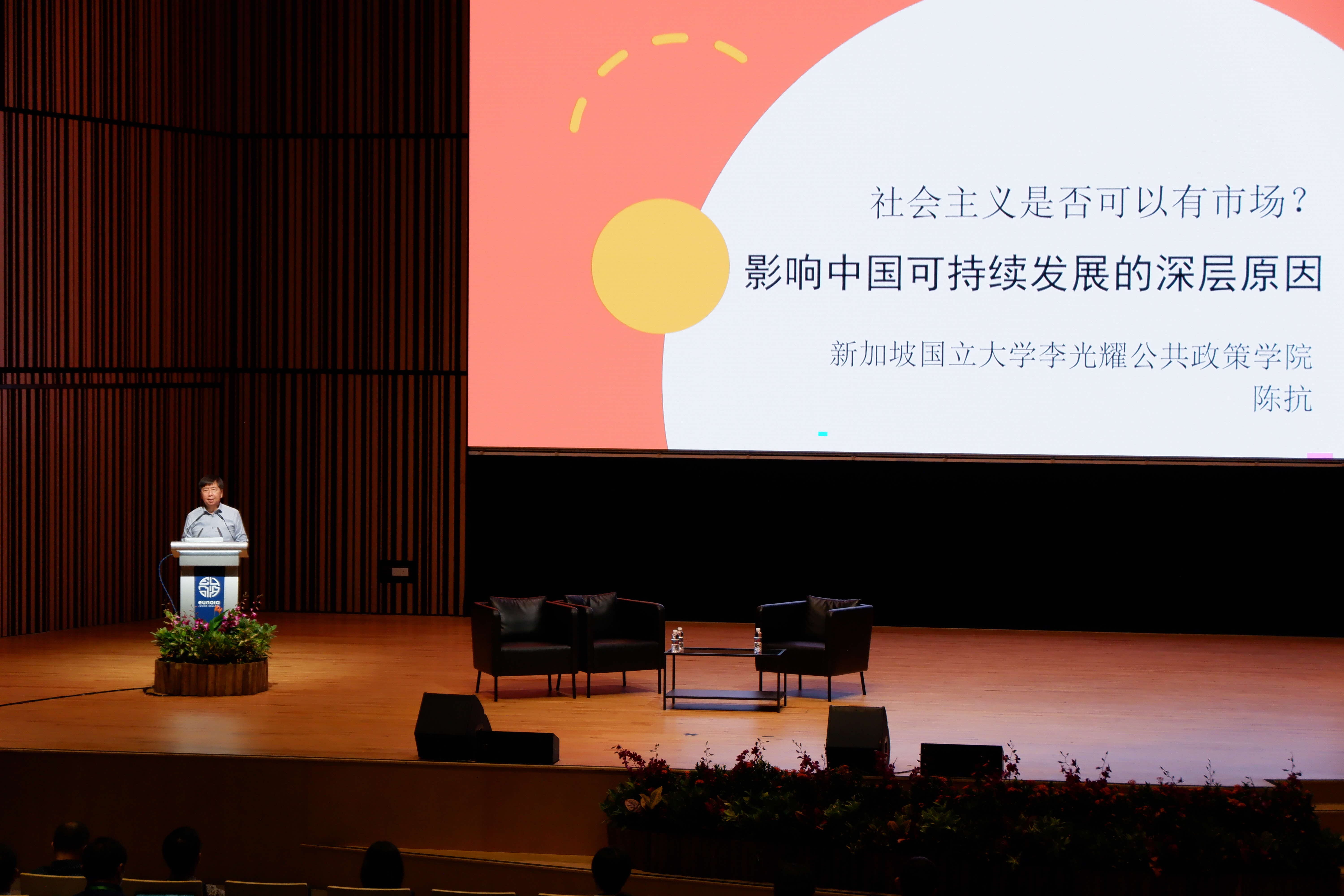 A person stands at a podium on a wooden stage with a large screen displaying Chinese text behind them.