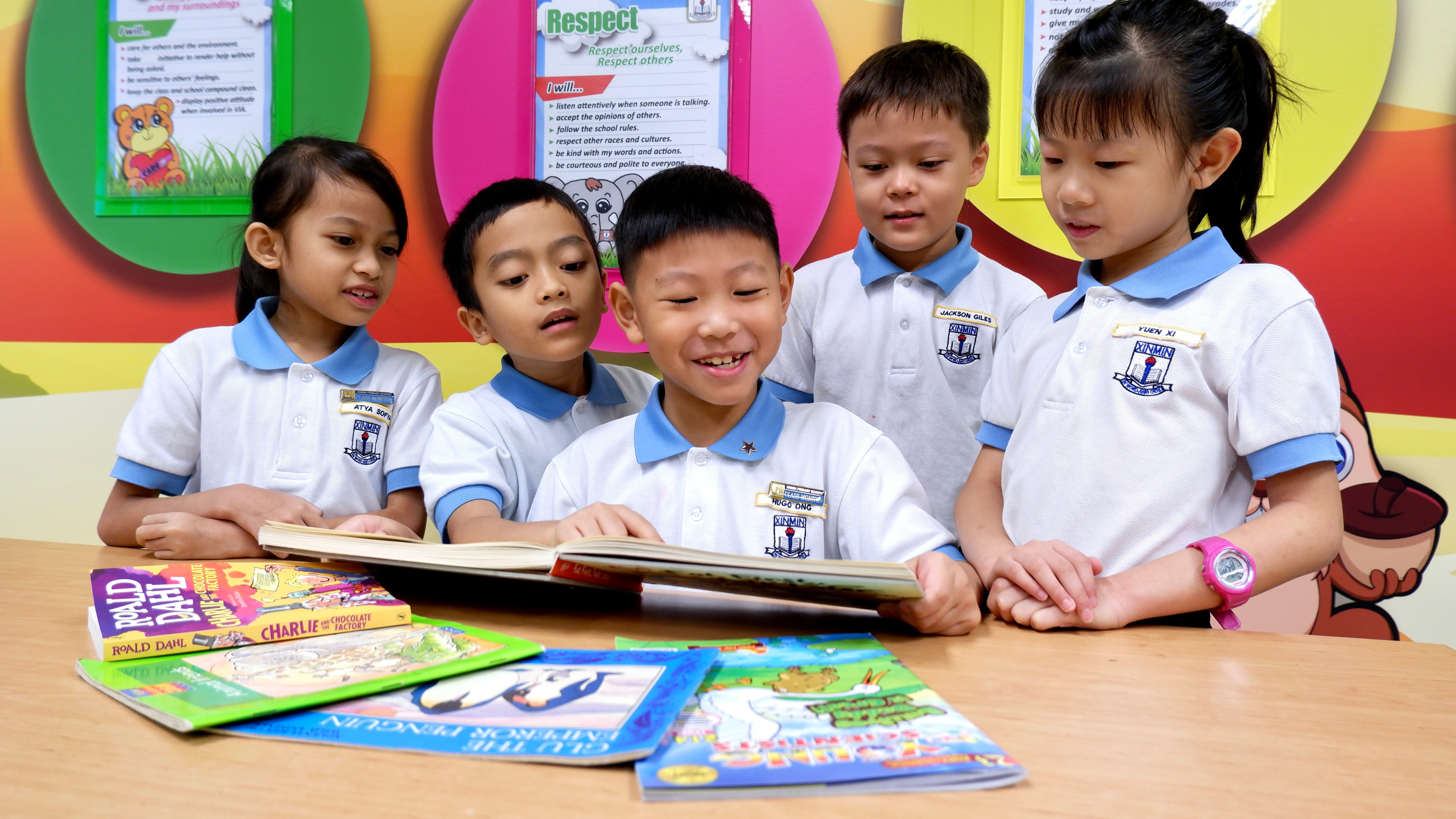 Five schoolchildren in white uniforms gather around a table, smiling at an open book.