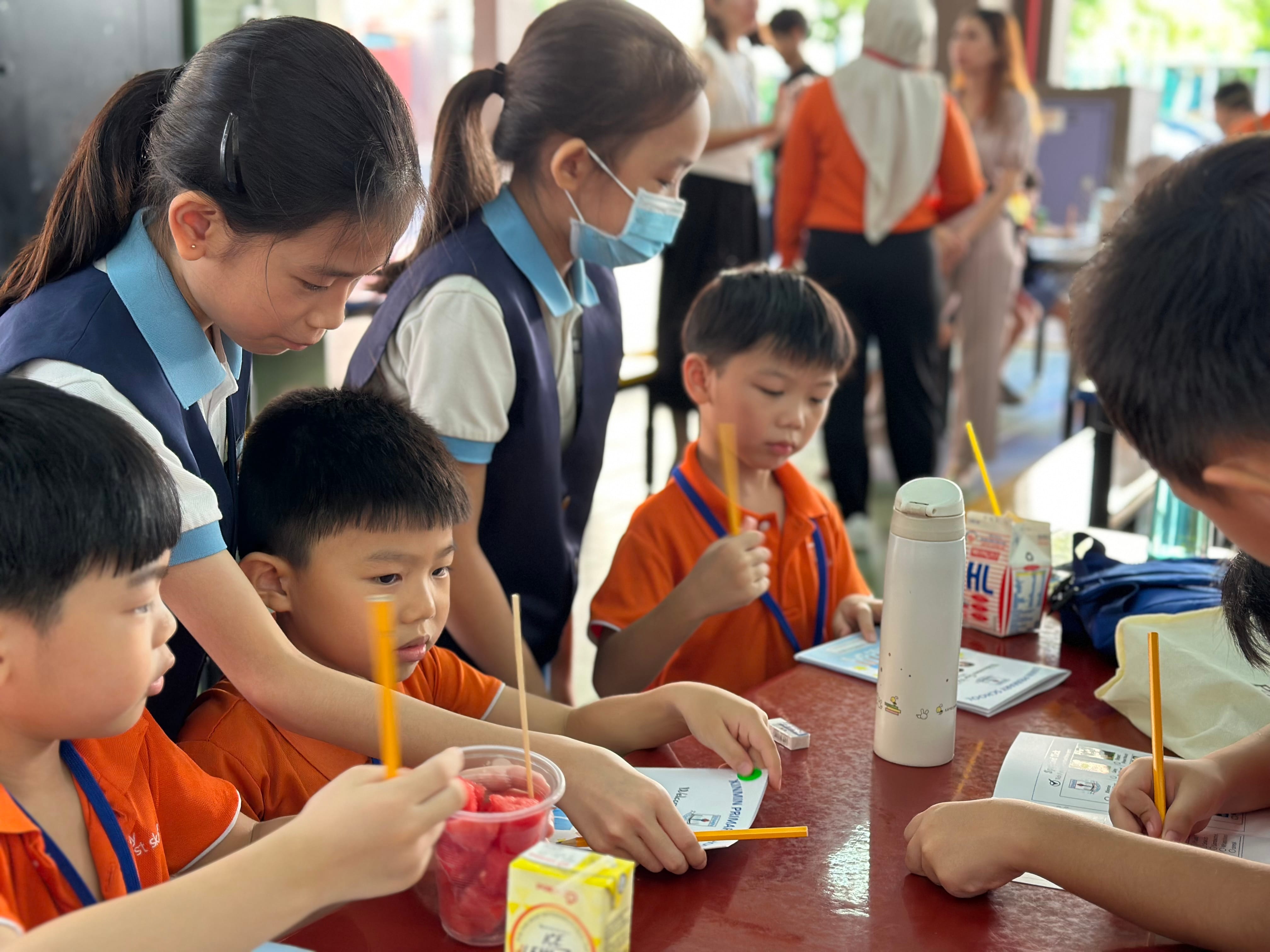 Young students in orange uniforms sit at a table with drinks and papers while older students in blue vests help them.
