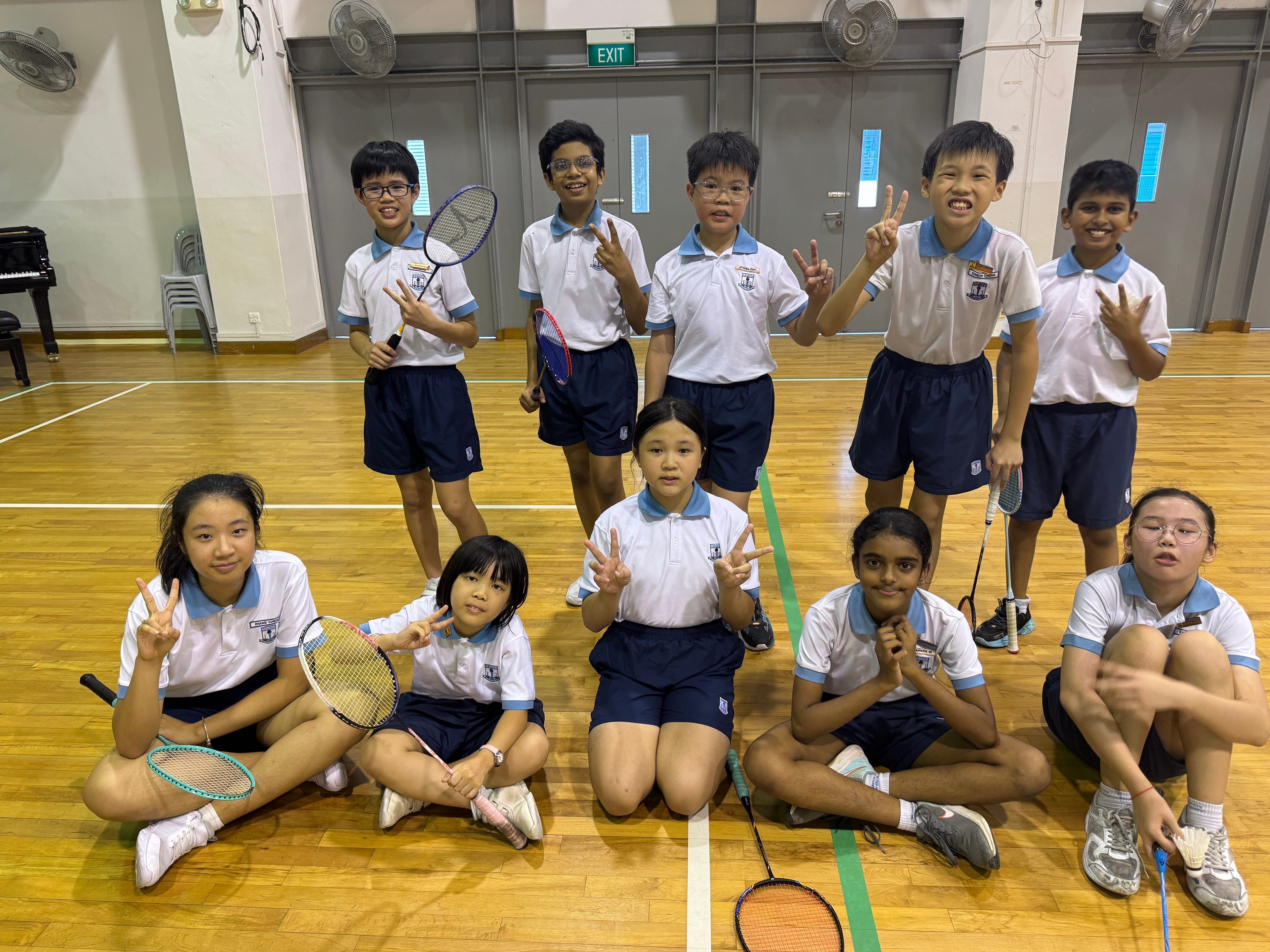 Ten school children in white and blue uniforms pose with badminton rackets on an indoor wooden court.