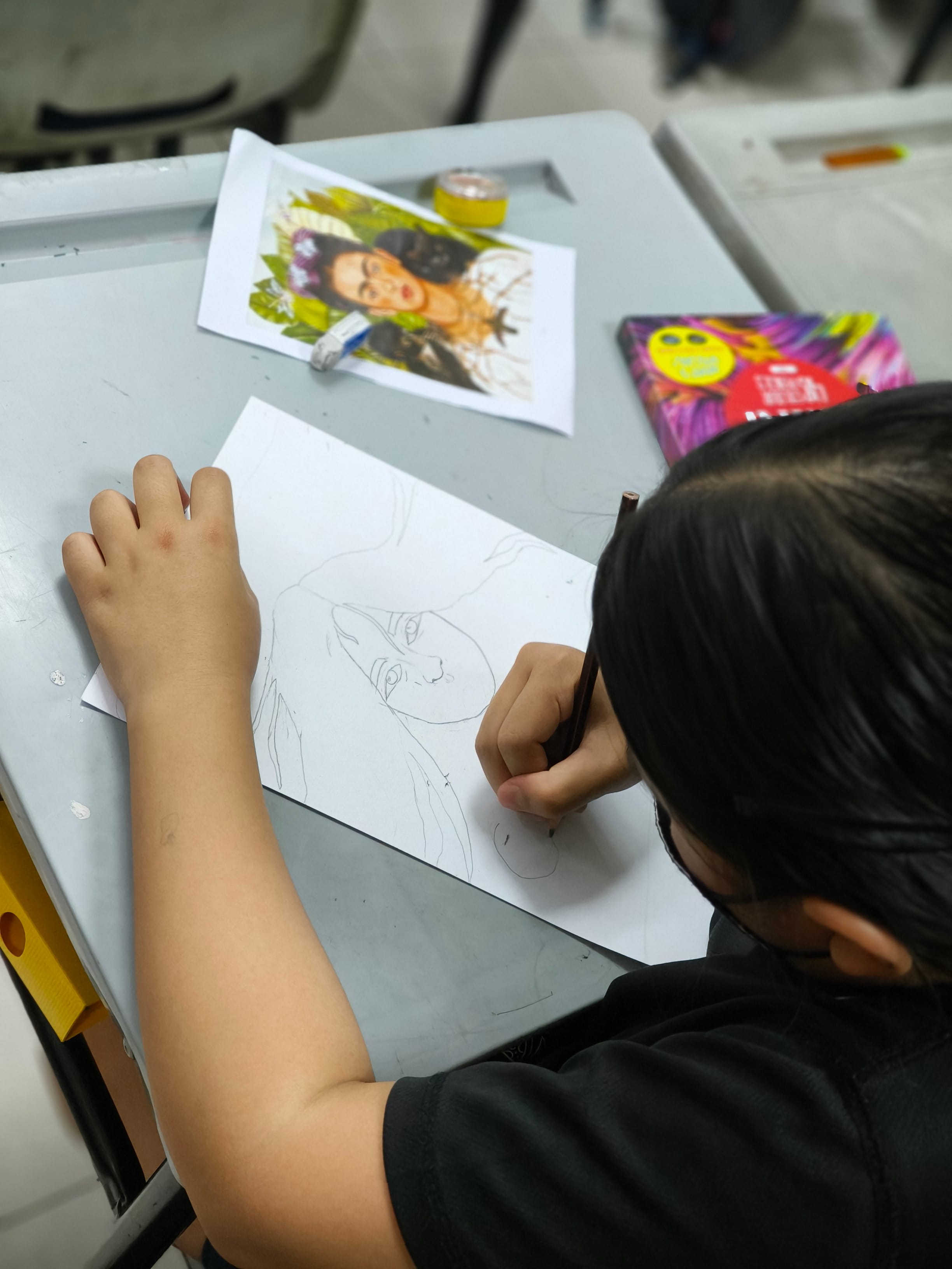 Child with dark hair sketching a portrait in pencil on paper, using a colorful reference photo of a woman on the desk.