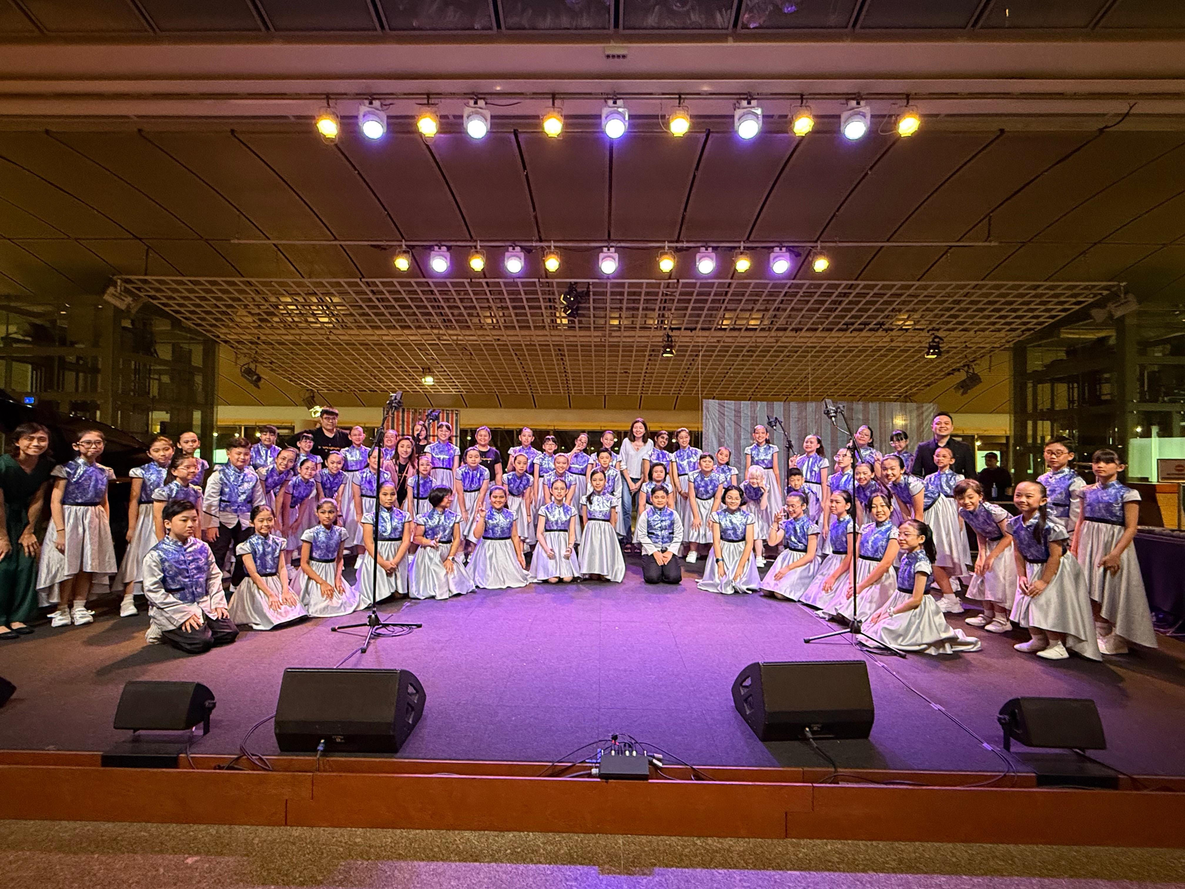 Large children's choir on a lit stage wearing matching blue and silver dresses, posing after a performance.