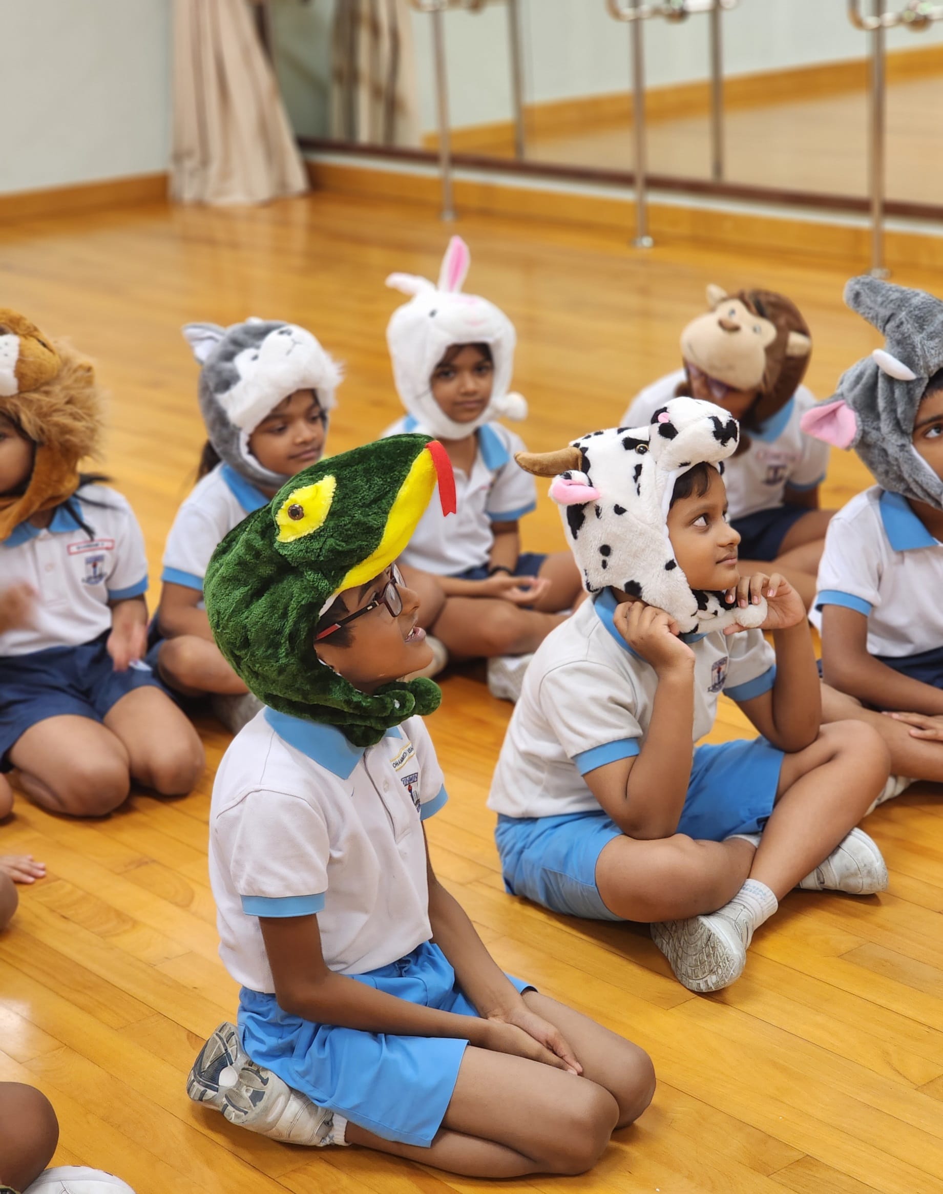 Children in school uniforms sit on a wooden floor wearing stuffed animal hats including a snake, cow, bunny, and monkey.
