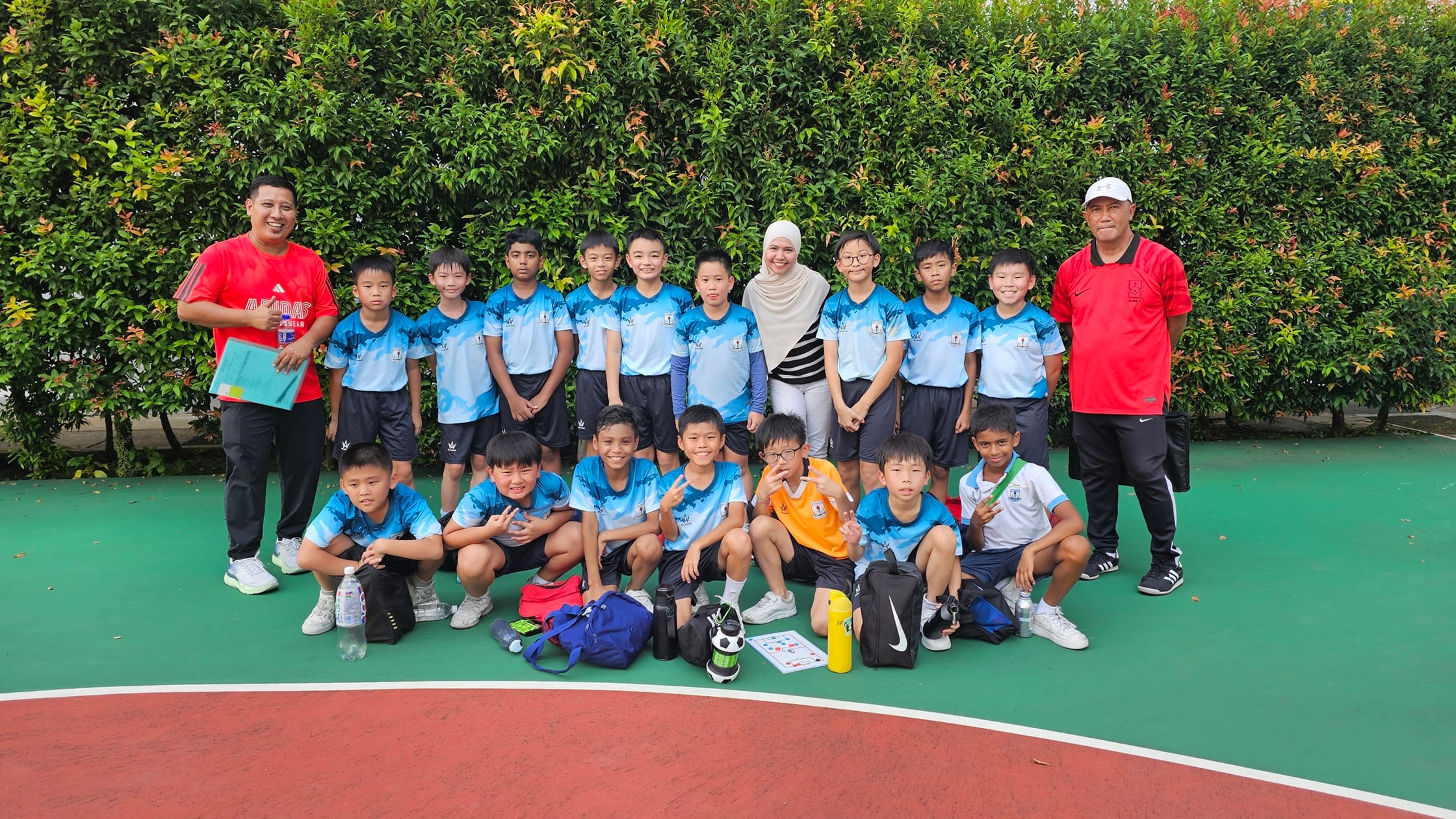 A youth soccer team in blue jerseys poses with two adult coaches on an outdoor sports court in front of green hedges.