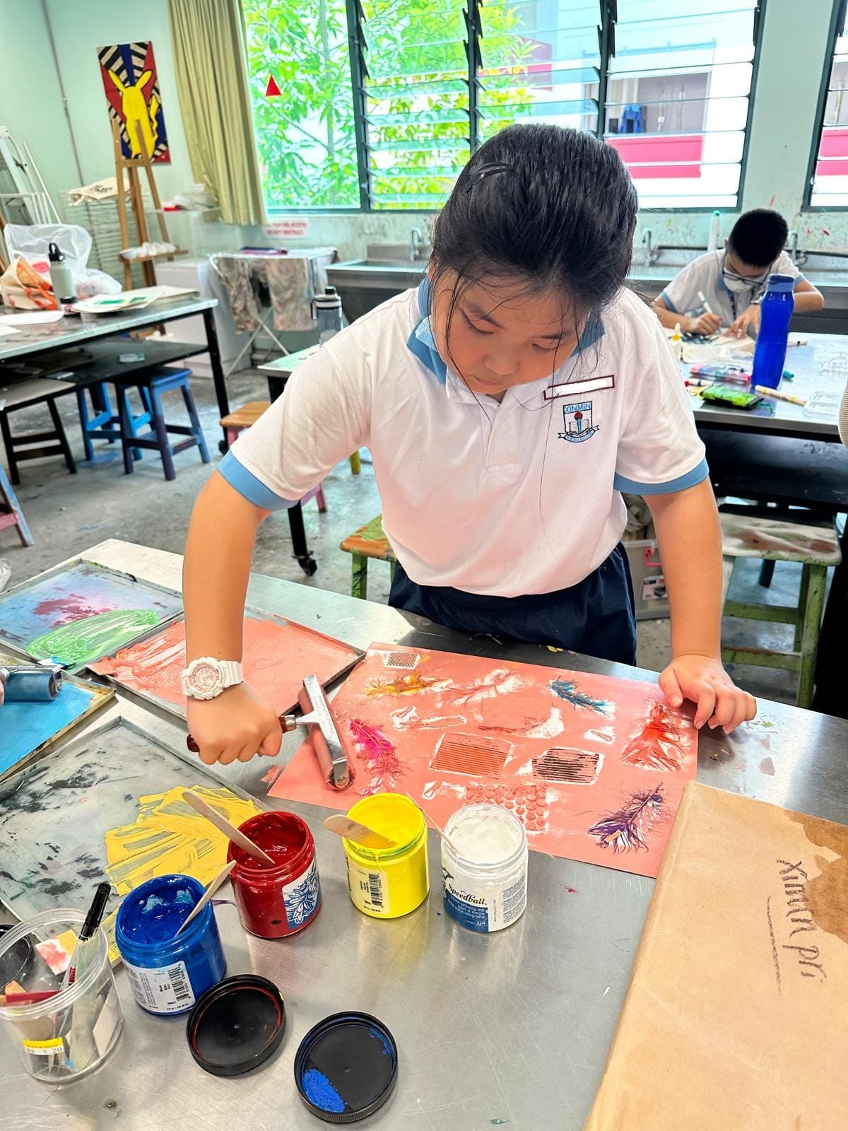 A student in school uniform works on a printmaking project at a table with jars of red, yellow, blue, and white paint.