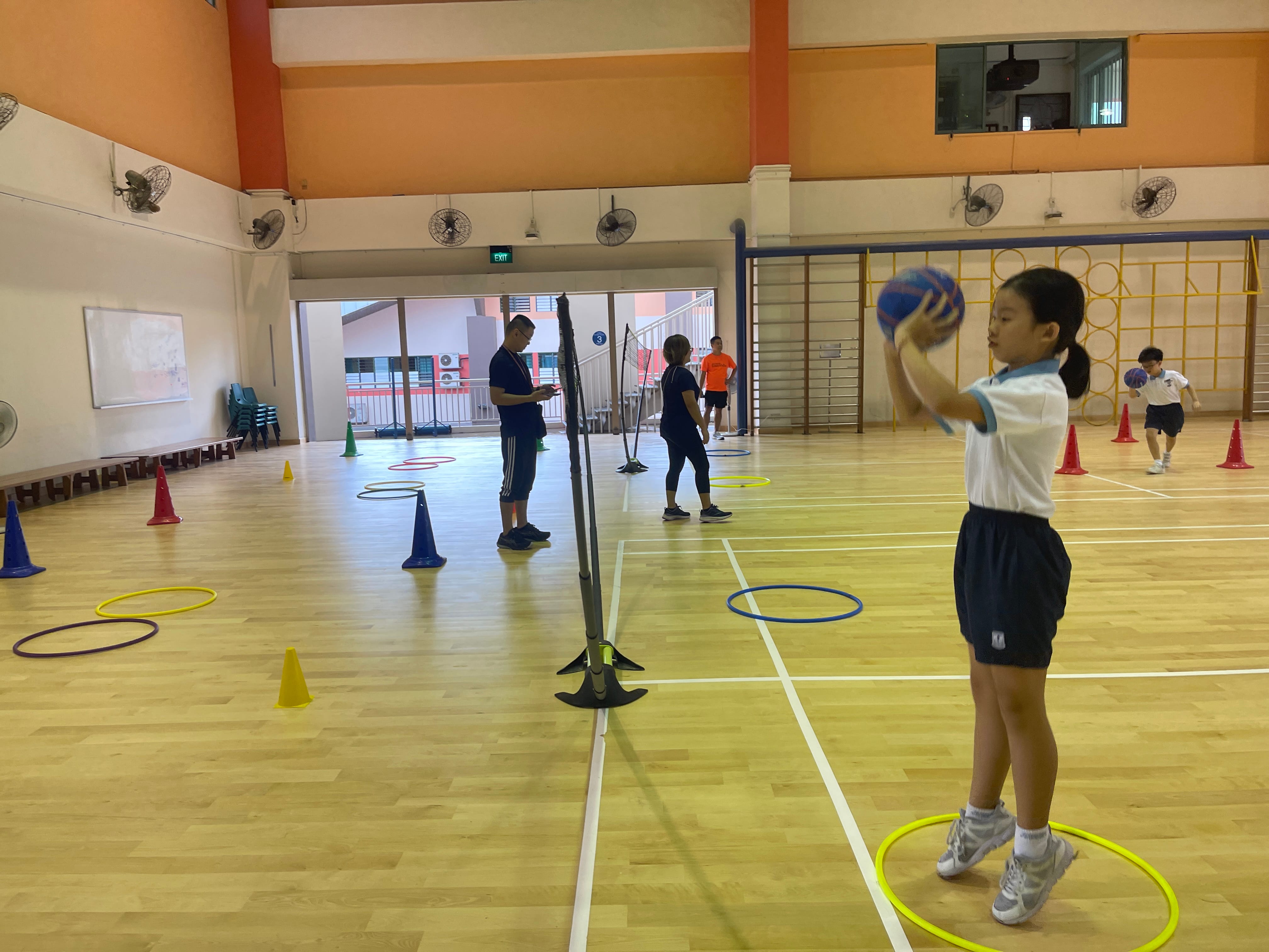 Children and adults practice ball skills in a school gymnasium with colorful hoops and cones on a wooden floor.