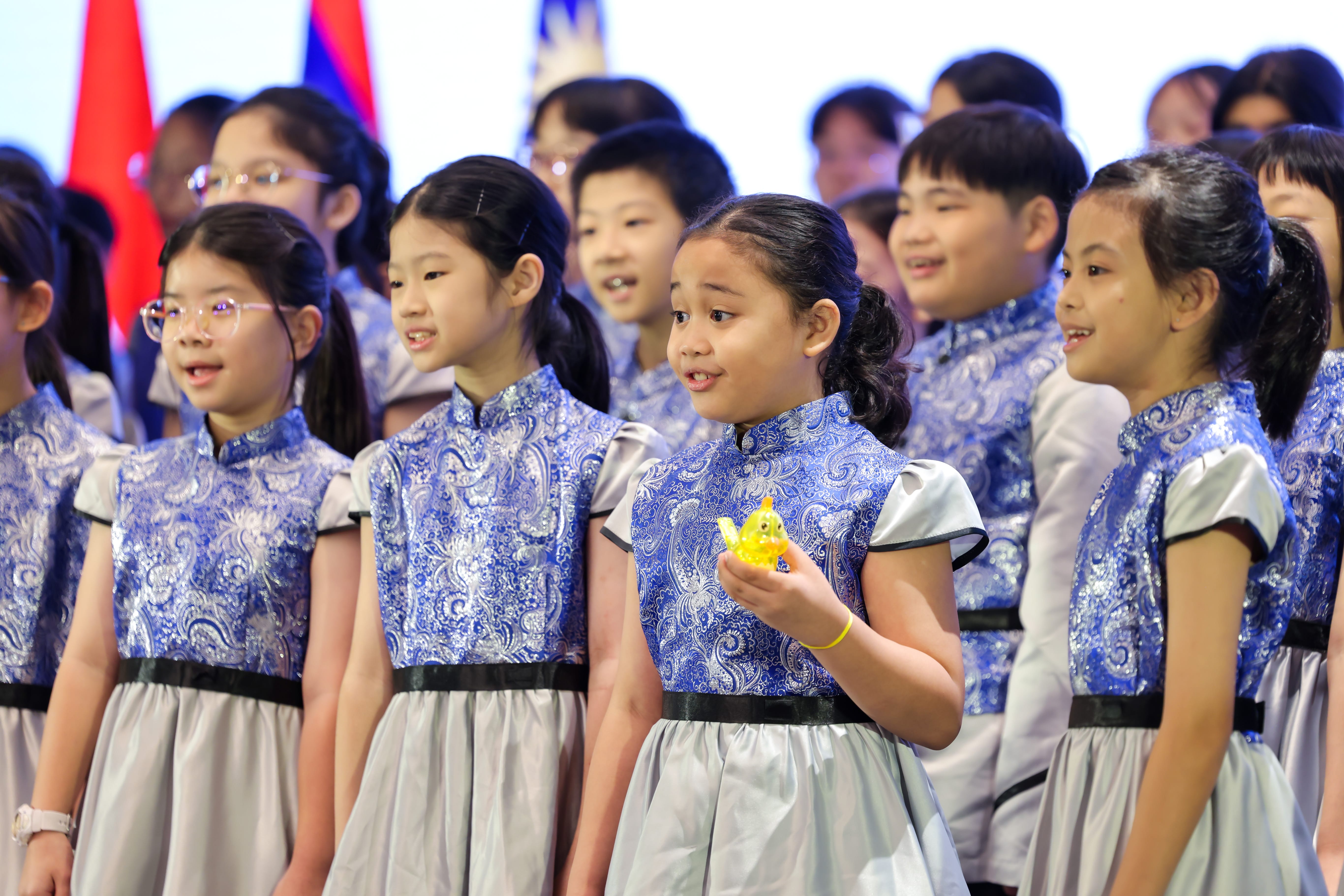 Children in matching blue paisley and silver dresses perform on stage, one girl holding a small yellow figurine.