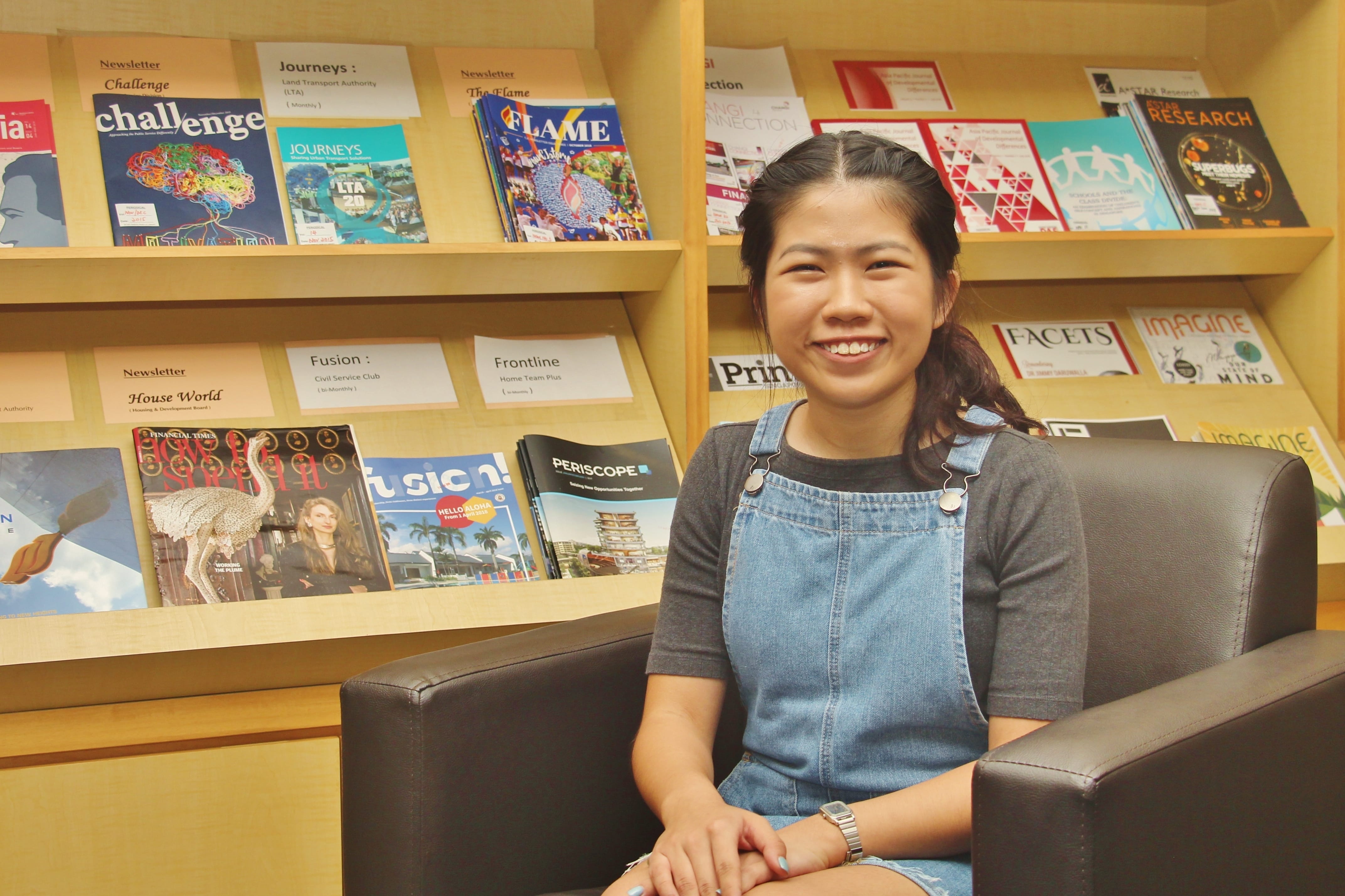 Smiling woman in denim overalls sits in a black armchair in front of wooden shelves of magazines.
