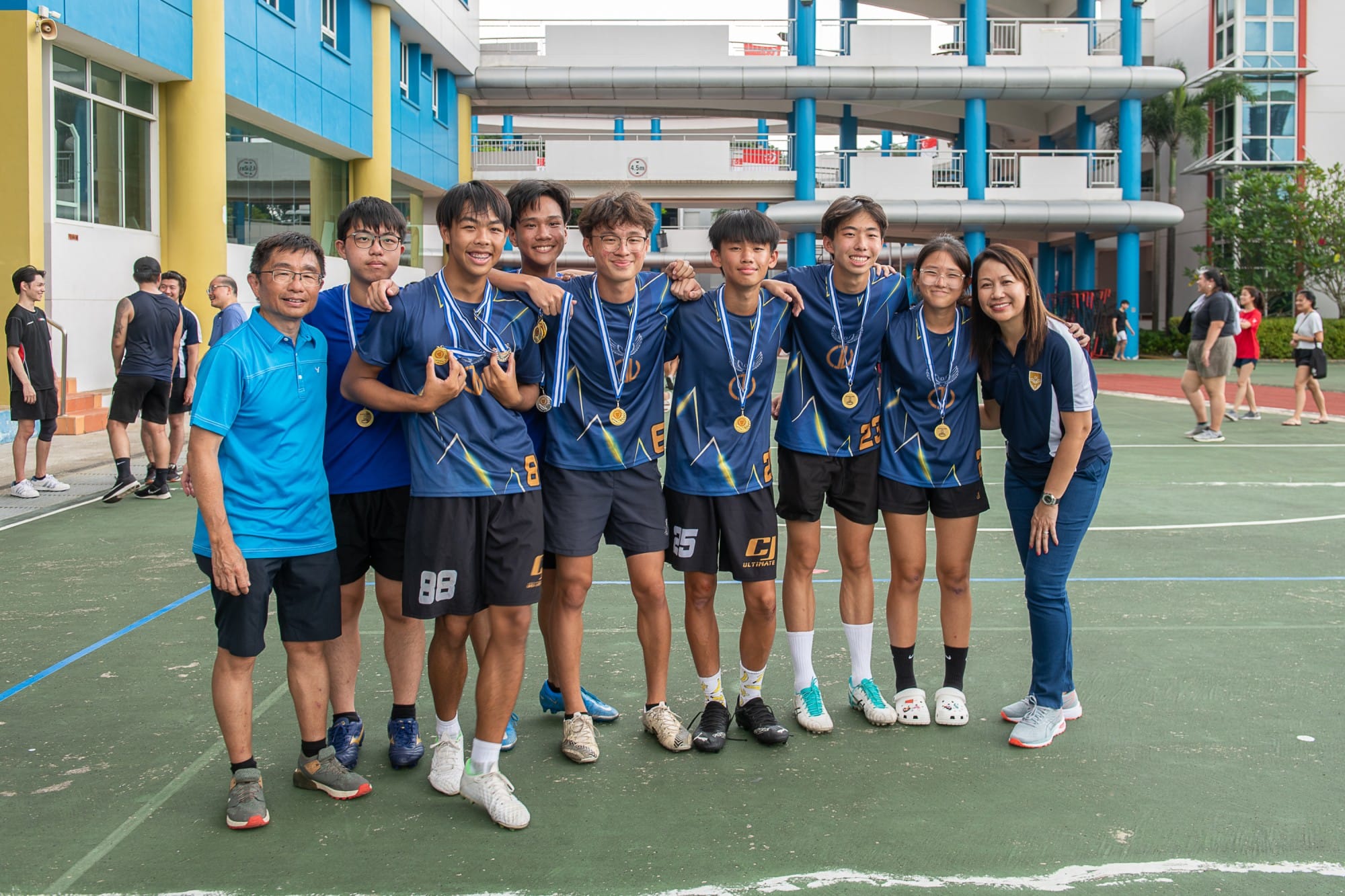 A group of young athletes in blue jerseys wearing gold medals pose with two coaches on an outdoor sports court.